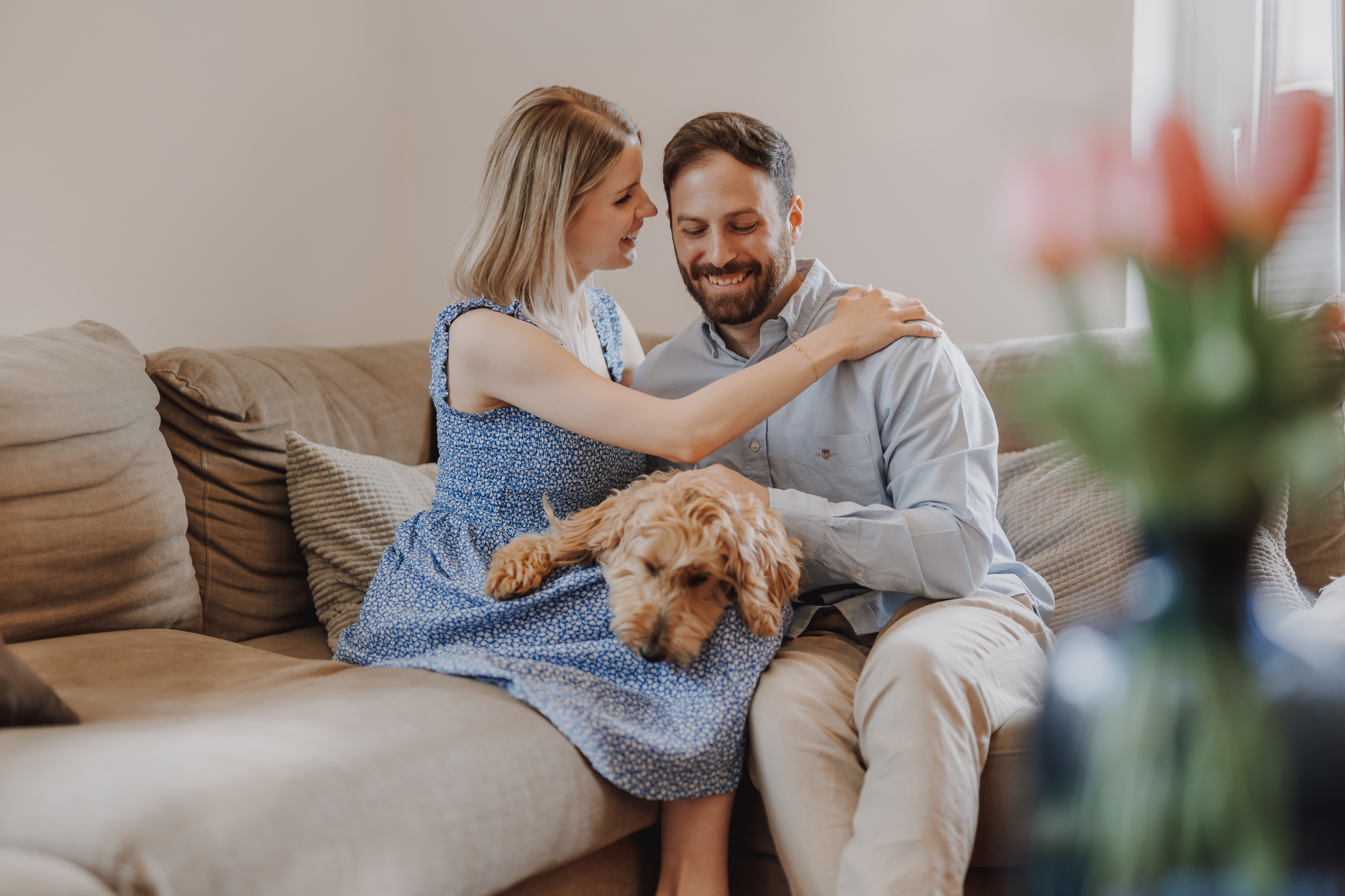 Eine Frau in einem blauen Kleid sitzt zu Hause auf einem Sofa, lächelt und berührt die Schulter eines Mannes in einem hellblauen Hemd neben ihr. Der Mann lächelt, während er einen braunen, lockigen Hund streichelt, der zwischen ihnen liegt. Im Vordergrund steht eine verschwommene Vase mit Blumen, die die Wärme ihres Schwangerschaftsshootings einfängt.