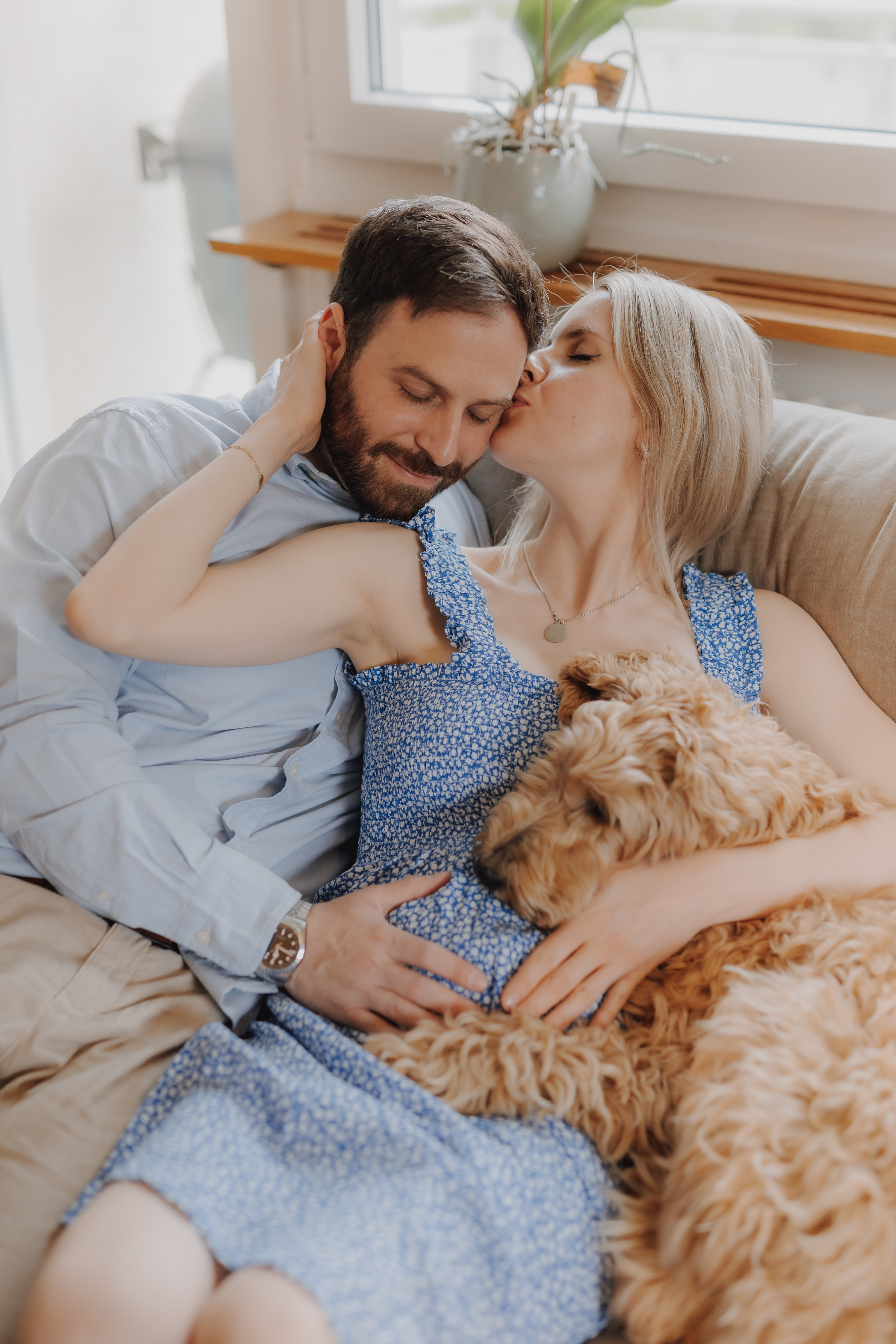Bei einem gemütlichen Babybauch-Fotoshooting küsst eine Frau in einem blauen Blumenkleid die Stirn eines bärtigen Mannes in einem hellblauen Hemd, während sie auf der Couch kuscheln. Mit ihrer Hand auf ihrem schwangeren Bauch und ihrem großen, lockigen Hund, der seinen Kopf auf ihrem Schoß ruht, fängt die Szene pure Glückseligkeit ein.
