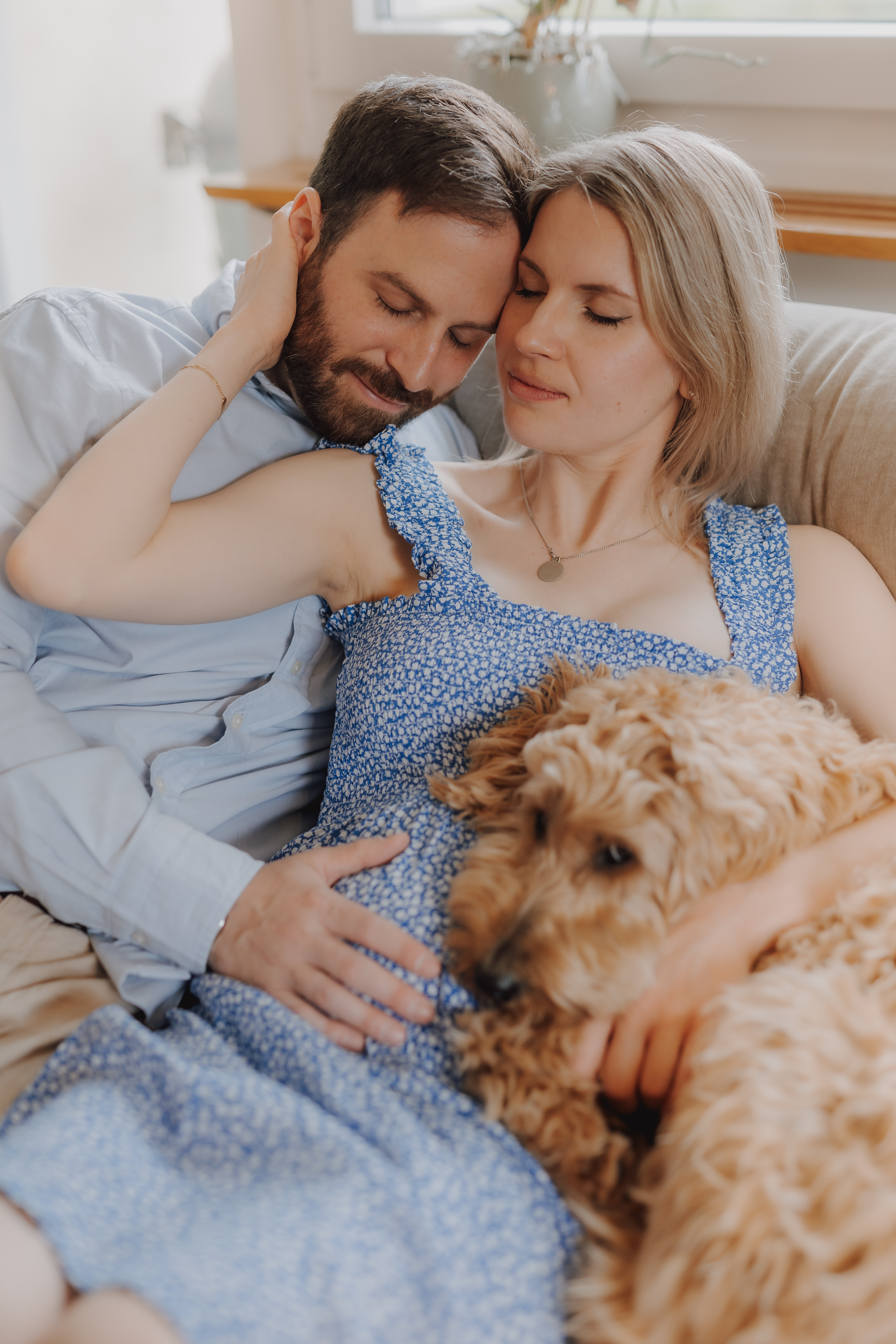 Ein Paar sitzt auf einer Couch und kuschelt sich aneinander und einen flauschigen braunen Hund. Der Mann trägt ein hellblaues Hemd und die Frau, die ein blaues Kleid mit Blumenmuster trägt, hat die Augen geschlossen und wirkt entspannt und glücklich während ihres Babybauch-Fotoshootings.