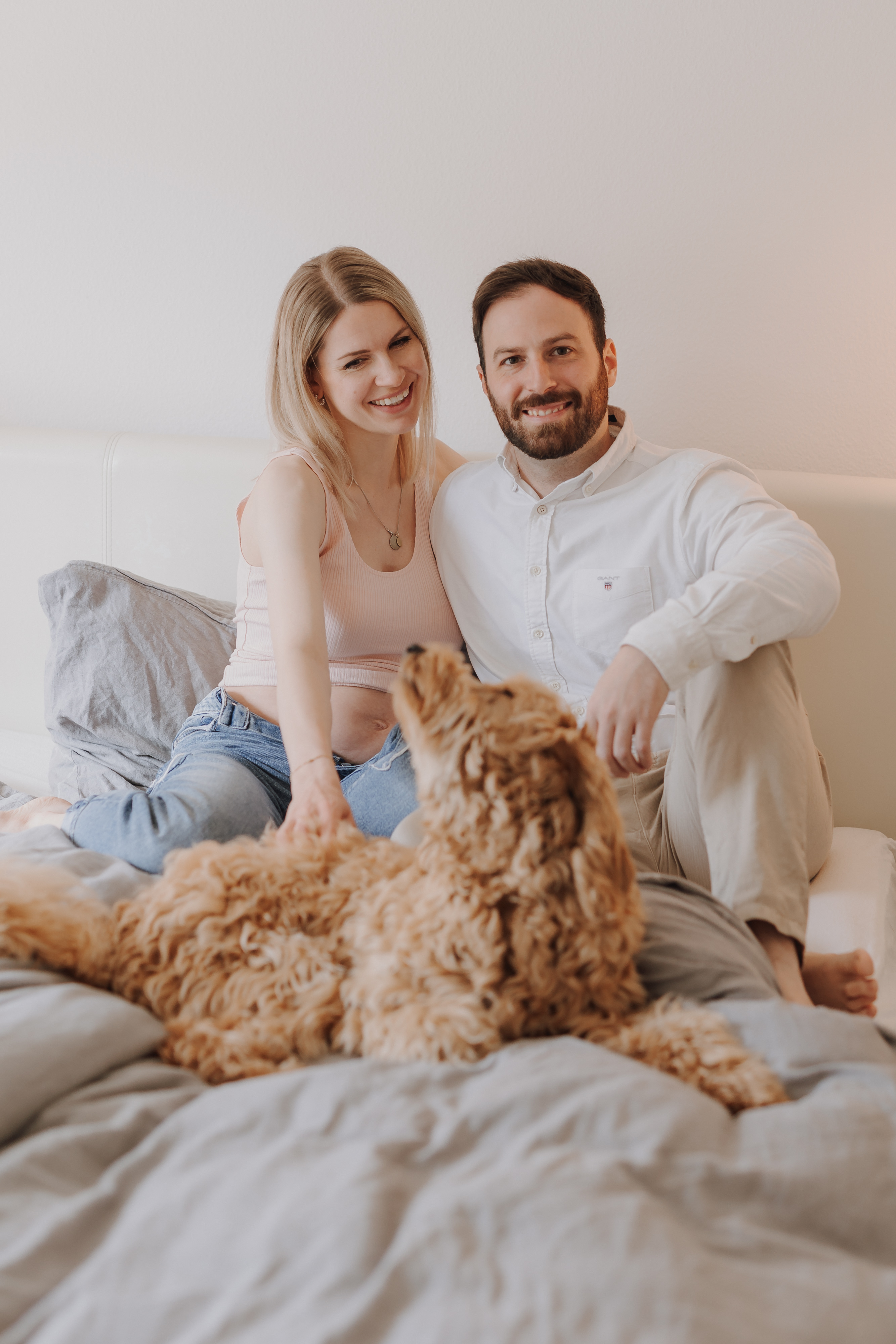 Ein lächelndes Paar sitzt auf einem Bett mit grau-weißer Bettwäsche, ein flauschiger Hund liegt gemütlich zwischen ihnen. Die Frau, die einen Babybauch in einem hellrosa Top und Jeans trägt, und der Mann in einem weißen Hemd und hellen Hosen wirken entspannt und glücklich.