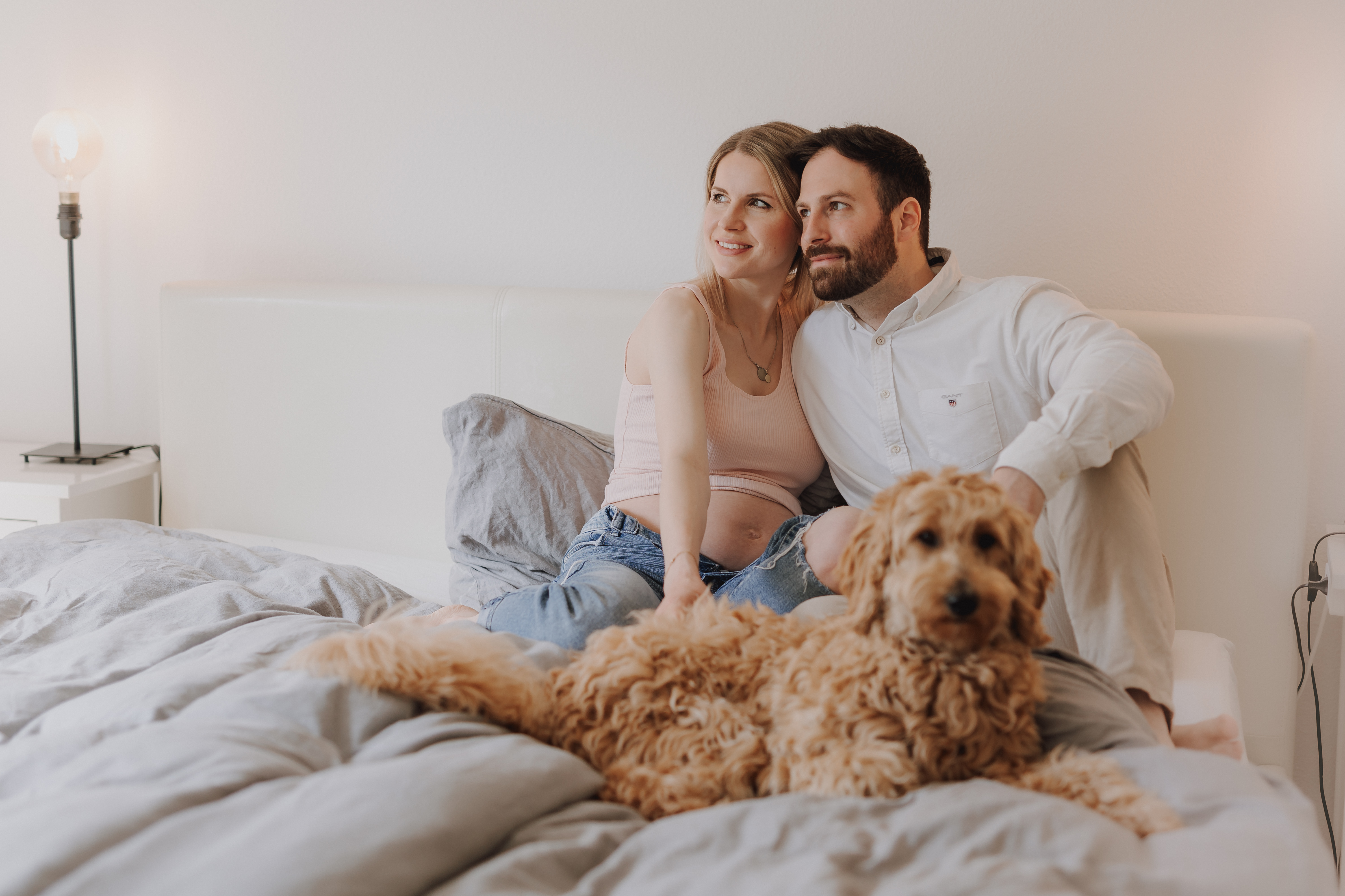 Ein Pärchen sitzt zufrieden auf einem Bett. Die Frau, die ein hellrosa Oberteil trägt, wiegt ihren Bauch und deutet damit auf ein Schwangerschafts-Fotoshooting hin, während der Mann neben ihr im weißen Hemd seinen Arm um sie legt. Im Vordergrund liegt ein flauschiger brauner Hund gemütlich auf dem Bett.