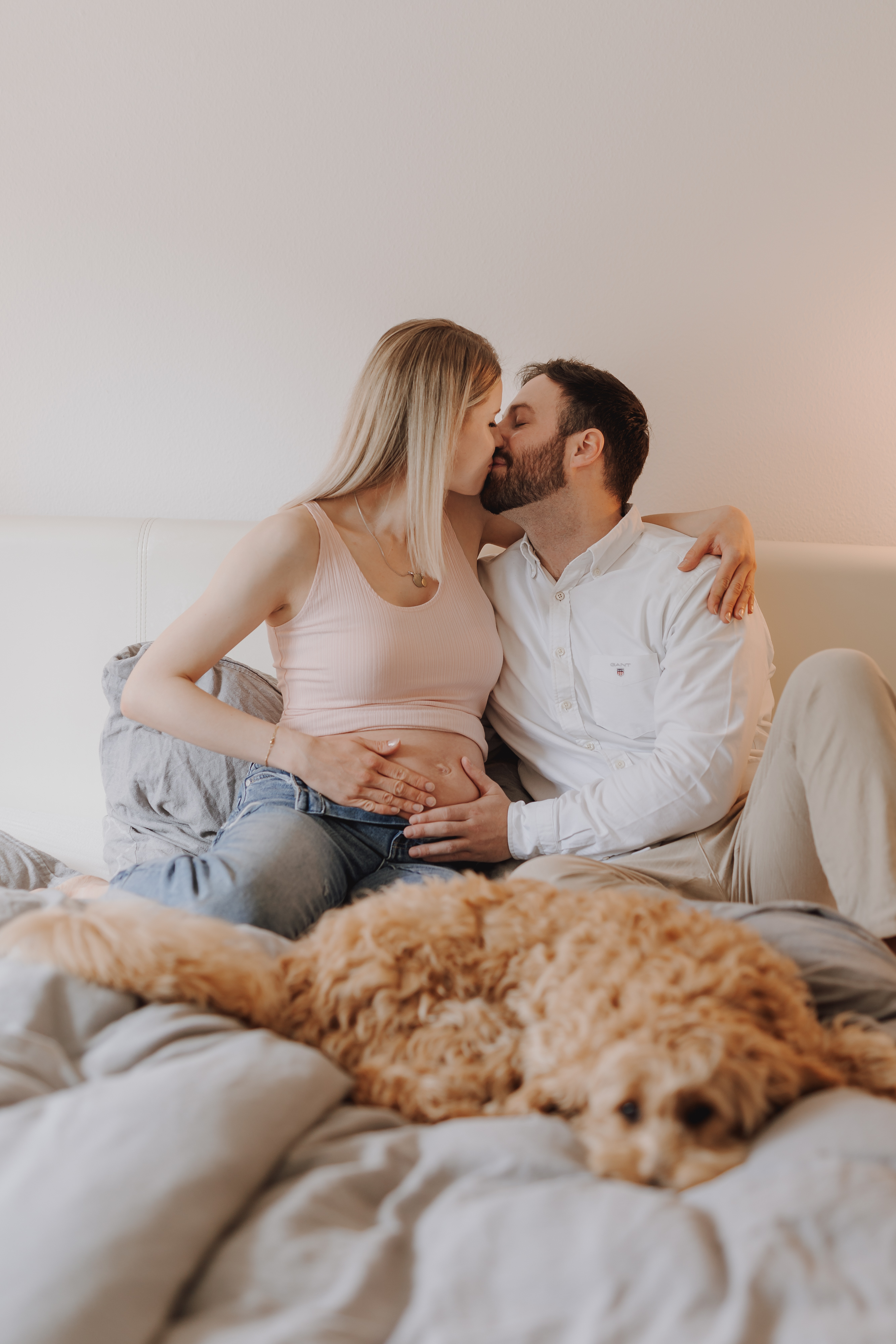 Eine schwangere Frau und ein Mann sitzen während ihres Schwangerschafts-Fotoshootings küssend auf einem Bett. Die Frau trägt ein rosa Tanktop und Jeans, die Hand des Mannes liegt auf ihrem Babybauch. Im Vordergrund liegt ein goldener Hund, der in die Kamera blickt. Das Paar ist umgeben von weicher Bettwäsche und einer gemütlichen Zuhause-Atmosphäre.