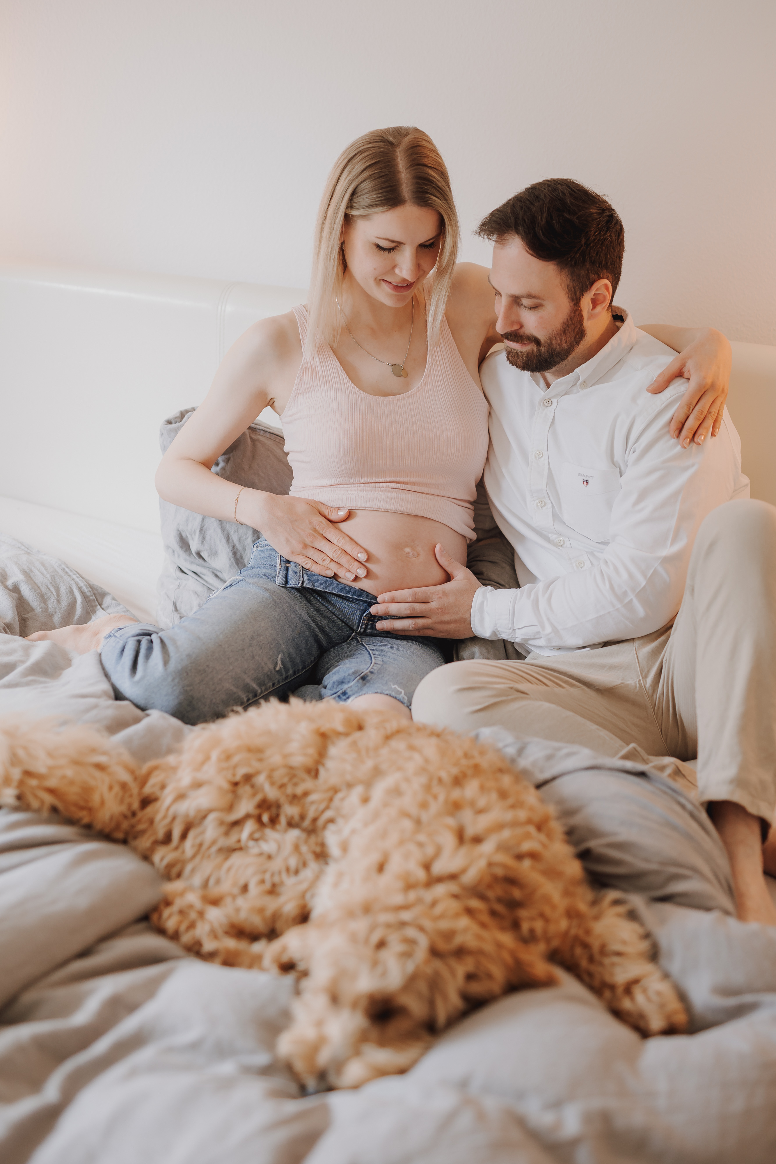 Eine schwangere Frau und ein Mann sitzen auf einem Bett, berühren liebevoll ihren Babybauch und lächeln während ihres Schwangerschafts-Fotoshootings. Sie werden von einem flauschigen Hund begleitet, der vor ihnen auf dem Bett liegt und zur gemütlichen und warmen Atmosphäre beiträgt.