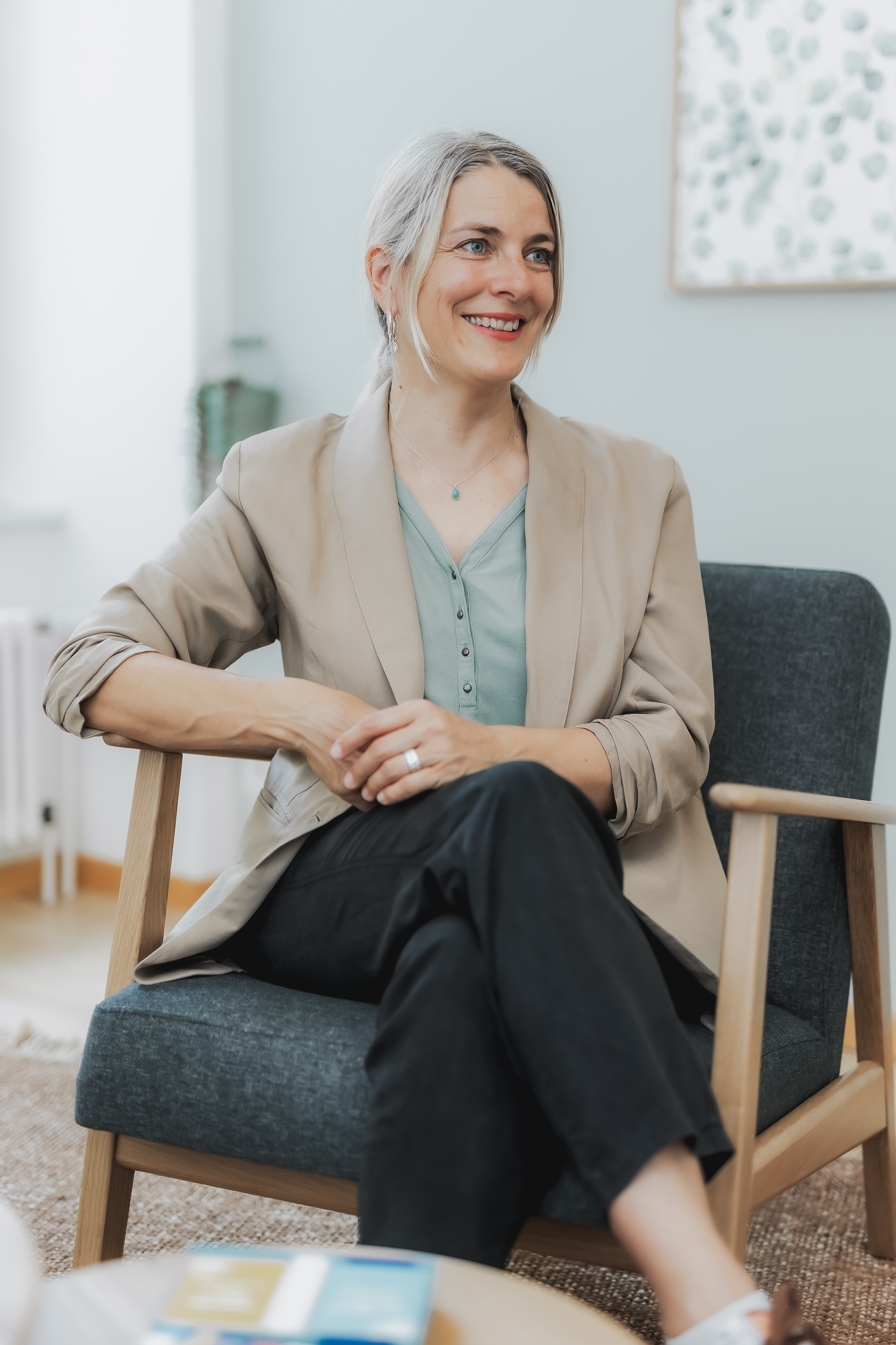 Eine Frau mit schulterlangem grauem Haar sitzt auf einem dunkelgrauen Sessel. Sie trägt einen beigen Blazer über einer blaugrünen Bluse und einer schwarzen Hose und lächelt, während sie die Beine übereinander schlägt und ihre Hände auf den Knien ruhen lässt. Der Hintergrund besteht aus einer Pflanze und Wandkunst und schafft die perfekte Szene für Content-Fotografie im eleganten Reportagestil.