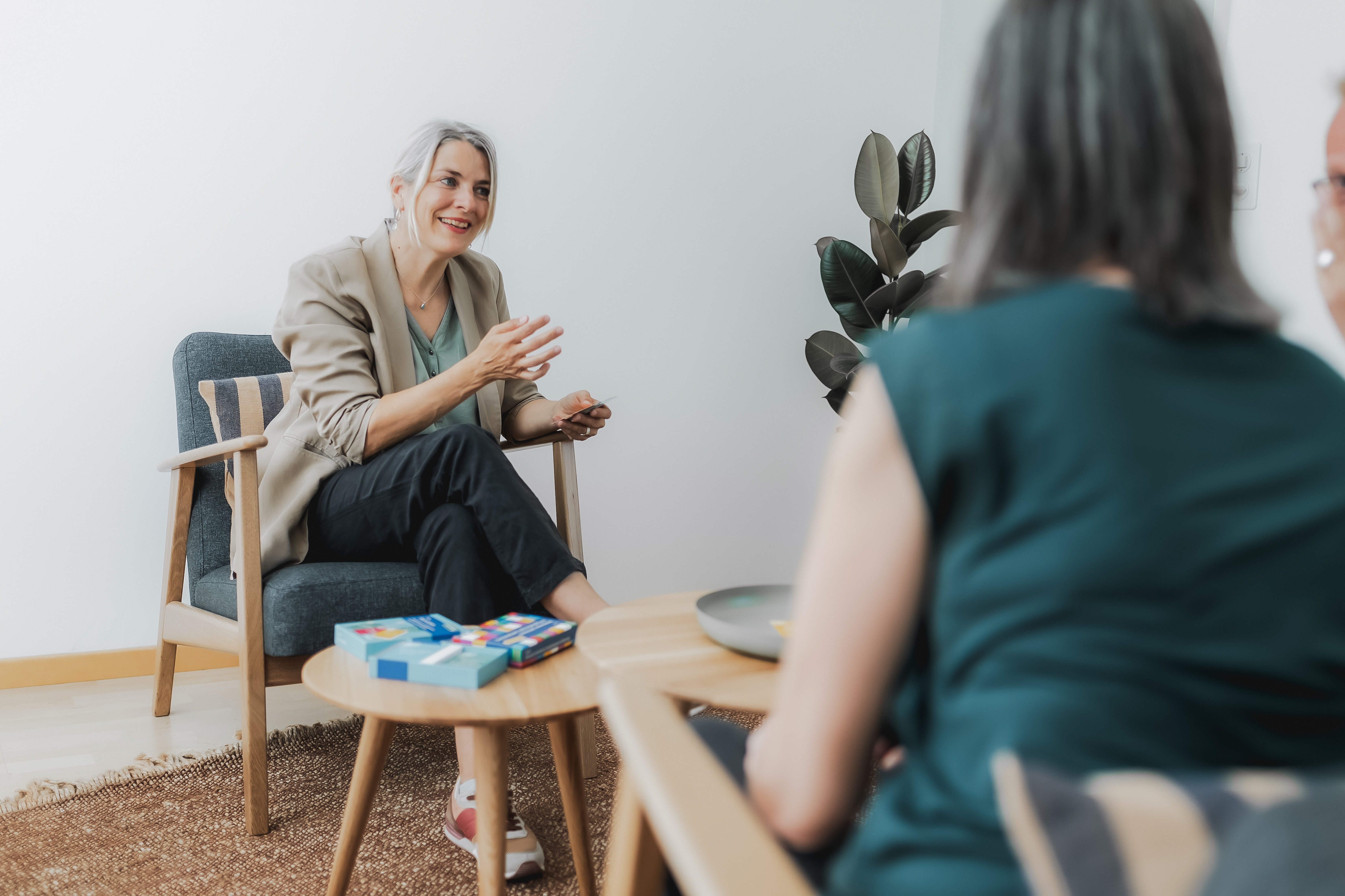 Eine Person mit grauem Haar lächelt und gestikuliert mit den Händen, während sie in einem Sessel sitzt und sich lebhaft unterhält, während sie an einer Firmenfotografie-Sitzung teilnimmt. Der gemütliche Raum mit hellen Wänden, Holzmöbeln und einem kleinen Tisch voller Kisten und Bücher bietet die perfekte Kulisse.
