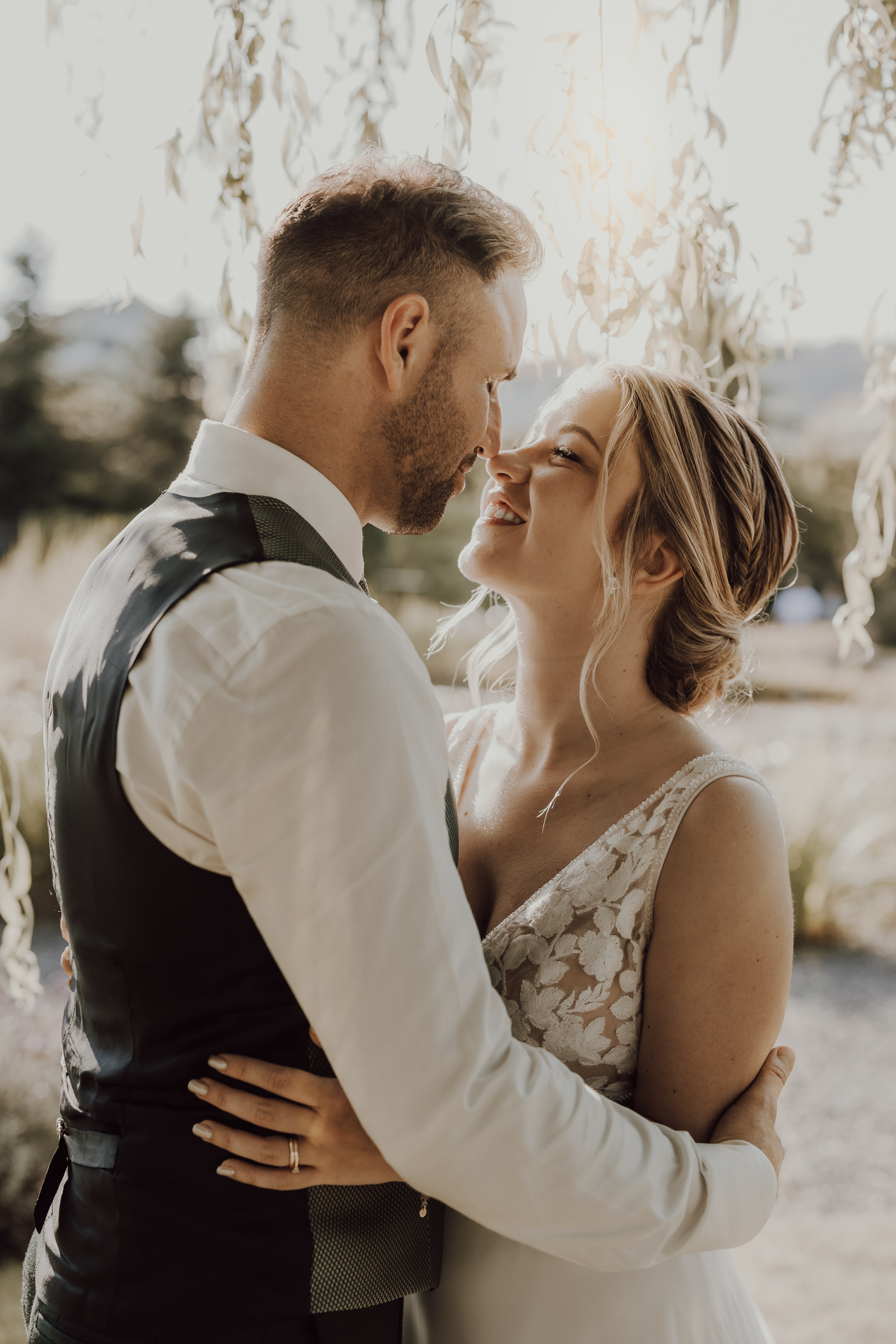 Ein Paar in Hochzeitskleidung genießt einen intimen Moment im Freien unter herabhängendem Laub. Der Bräutigam trägt ein weißes Hemd und eine Weste, während die Braut ein Spitzenhochzeitskleid trägt. Hochzeitsfotografin Tasha Ender fängt ihre Liebe ein, als sie sich liebevoll anschauen, während sanftes Sonnenlicht durch die Blätter fällt.