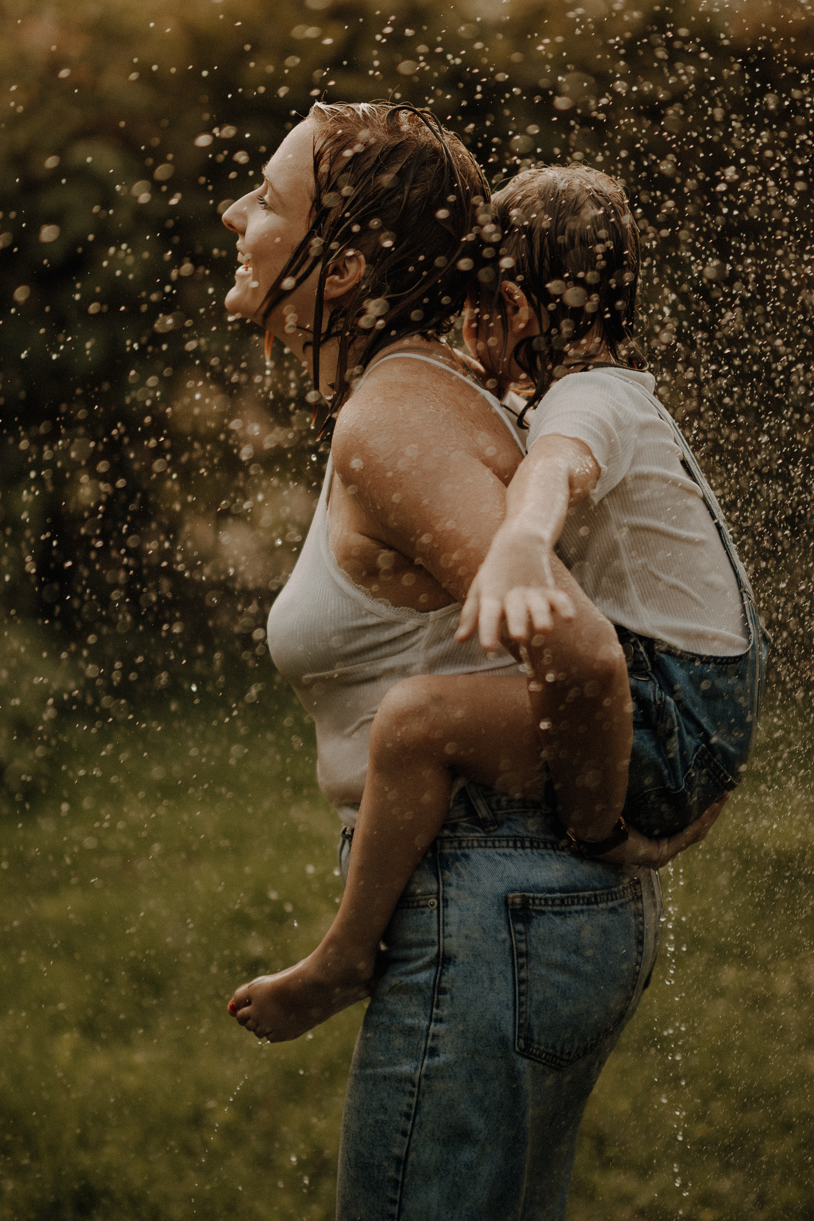Eine Frau mit nassen Haaren, einem weißen Tanktop und Jeans trägt ein kleines Kind auf dem Rücken. Das ebenfalls nasse Kind streckt spielerisch die Arme aus. Beide scheinen einen Regenschauer inmitten einer grünen Außenkulisse zu genießen, was den perfekten Moment für ein spontanes Familienshooting schafft.