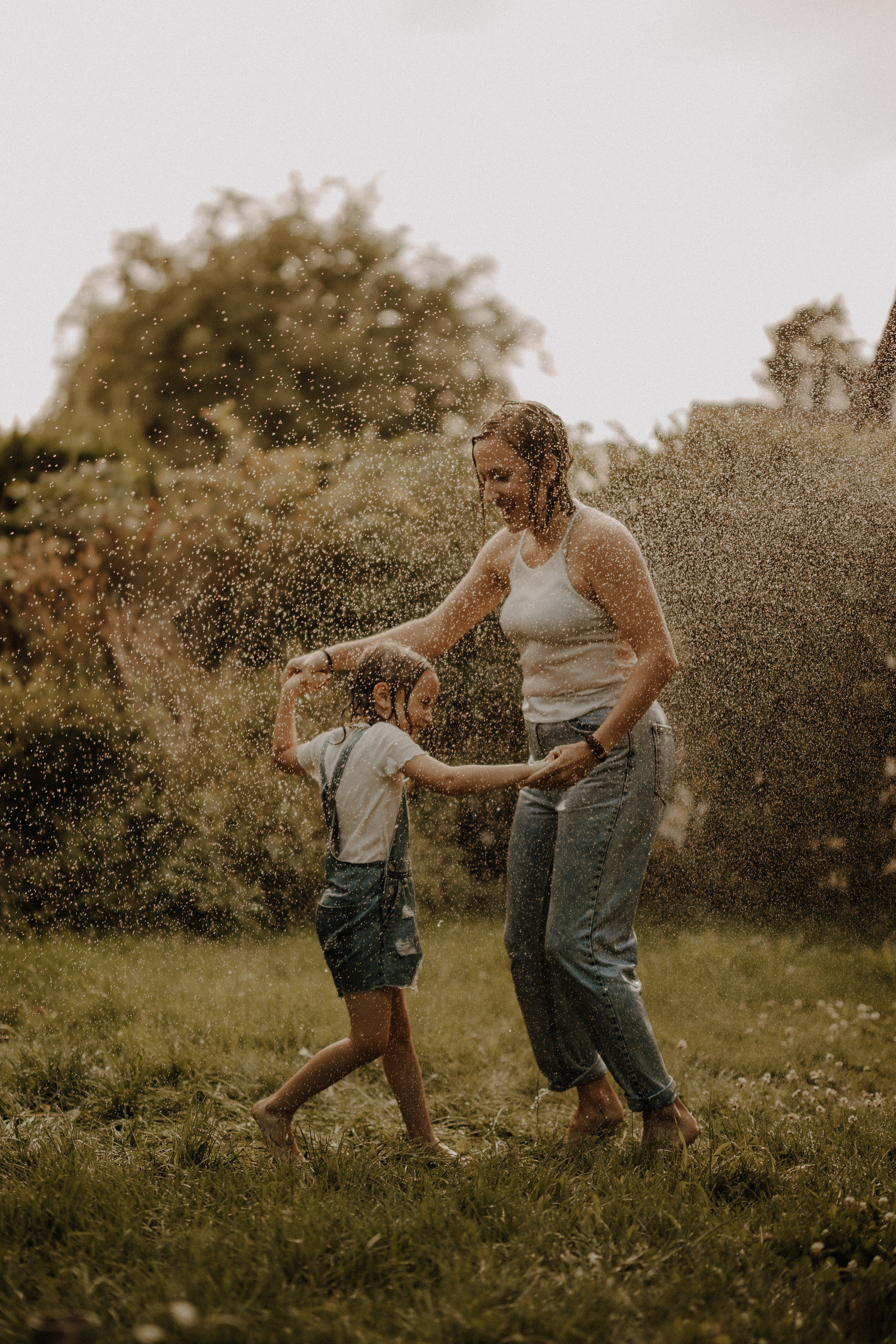Eine Frau und ein Kind spielen fröhlich in einem Garten während eines Muttertags-Fotoshootings und laufen an einem sonnigen Tag durch eine Wasserfontäne. Die Frau trägt ein weißes Tanktop und Jeans, während das Kind ein weißes T-Shirt und einen Overall trägt. Beide lächeln und scheinen Spaß zu haben.