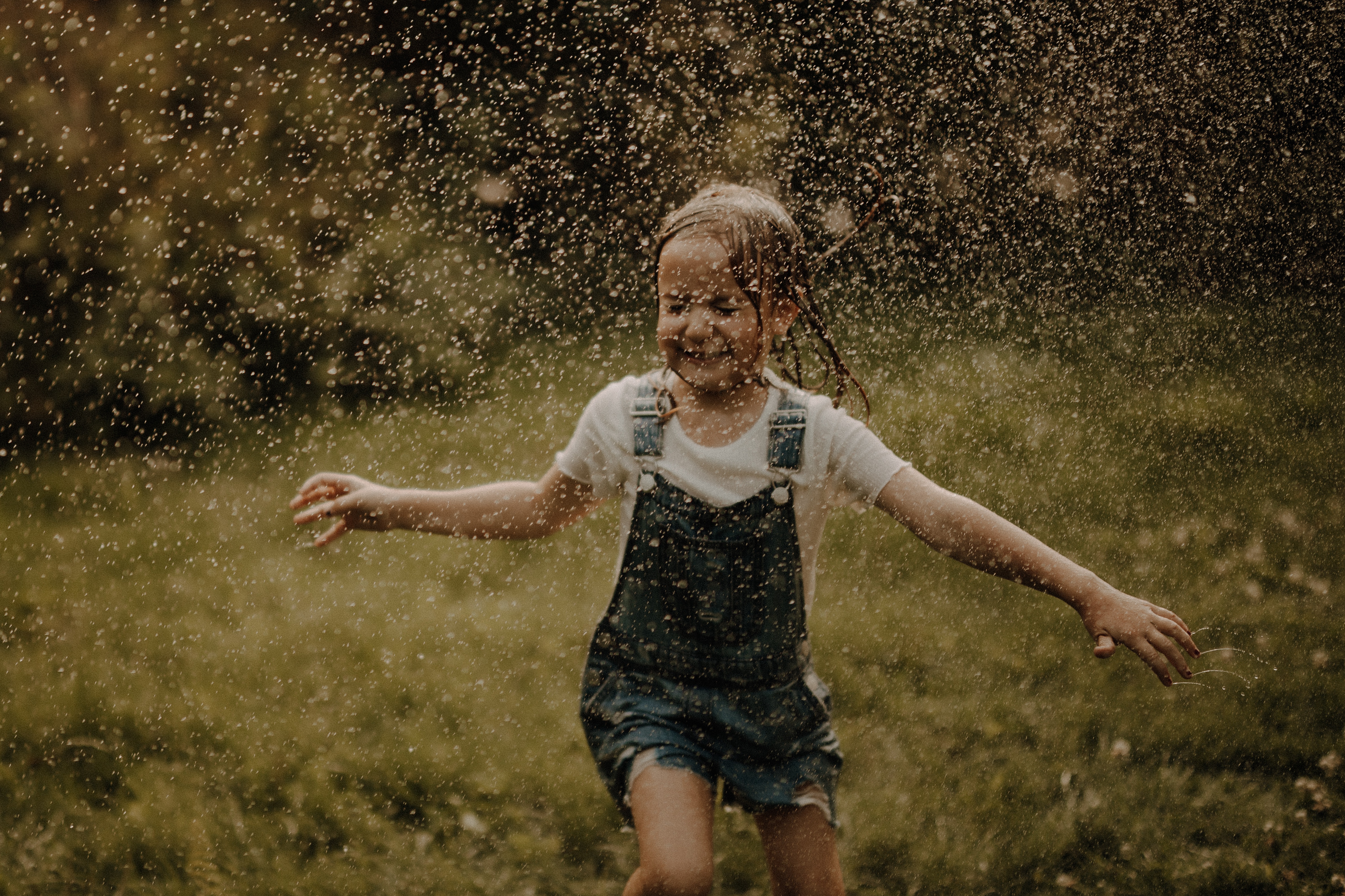 Ein junges Mädchen in Jeansoverall und weißem Hemd läuft fröhlich durch eine Gischt aus Wassertropfen auf einer Wiese vor einem grünen Hintergrund. Sie hat nasse Haare und ein breites Lächeln und fängt den verspielten Moment während des Familienshootings ein.
