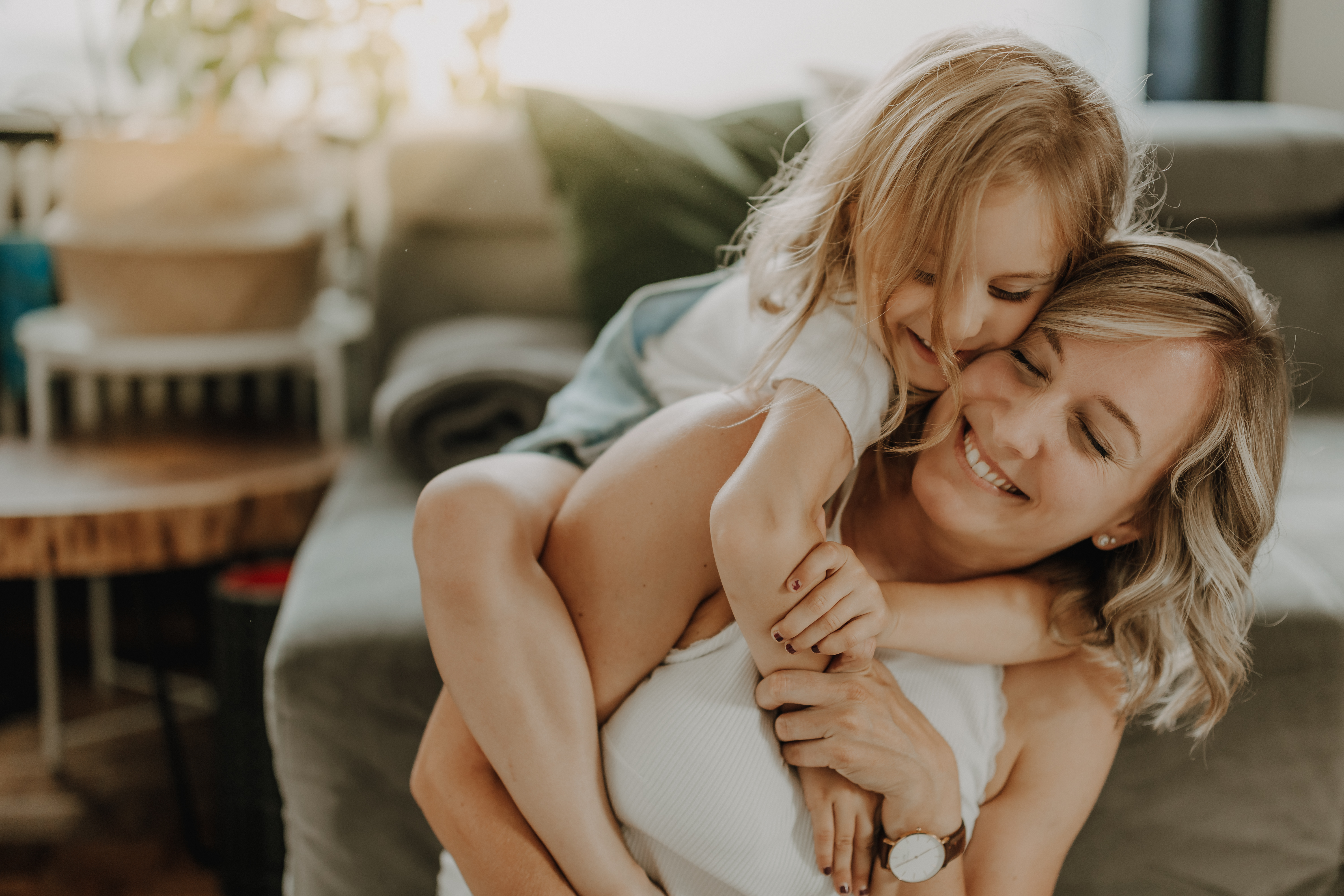 In einem gemütlichen Wohnzimmer in Baden, Aargau, trägt eine lächelnde blonde Frau ihre junge blonde Tochter huckepack im Rahmen des Fotoshootings von Tasha Ender Photography. Das junge Mädchen umarmt ihre Mutter freudig von hinten. Im Hintergrund sind ein Sofa, ein Holztisch und sanftes Tageslicht zu sehen, das durch ein Fenster fällt.
