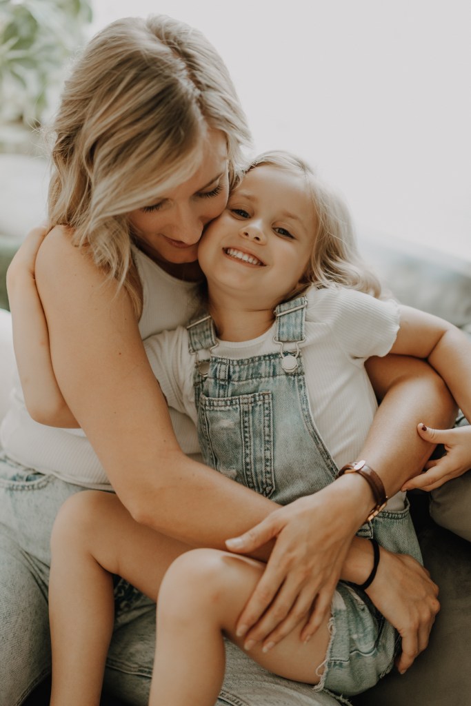 Eine Frau mit blonden Haaren umarmt ein fröhliches Kind, das in Jeans-Overall und weißem Hemd gekleidet ist. Sie sitzen auf einem Sofa in einem hell erleuchteten Raum, wirken fröhlich und entspannt und genießen ihre Momente während eines Fotoshootings von Tasha Ender Photography bei Zuhause.