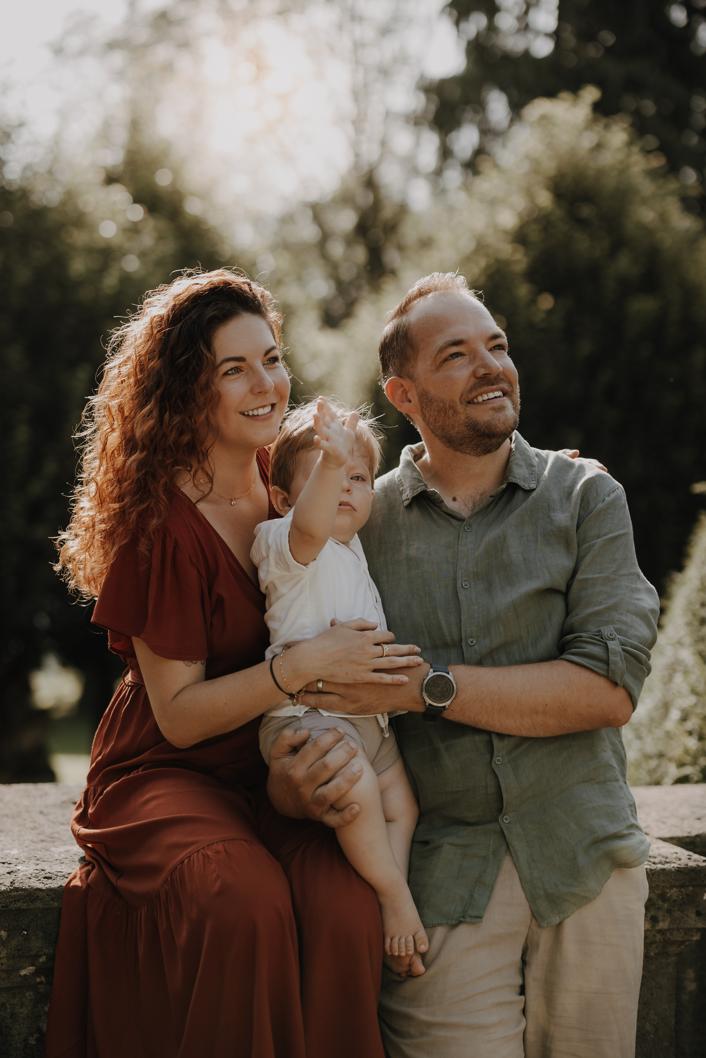 Eine Frau im roten Kleid und ein Mann im grünen Hemd stehen dicht beieinander, beide lächeln und blicken in die Ferne während ihres Fotoshootings in der Natur. Die Frau hält ein kleines Kind in einem weißen Outfit, das nach oben zeigt, umgeben von üppigem Grün.