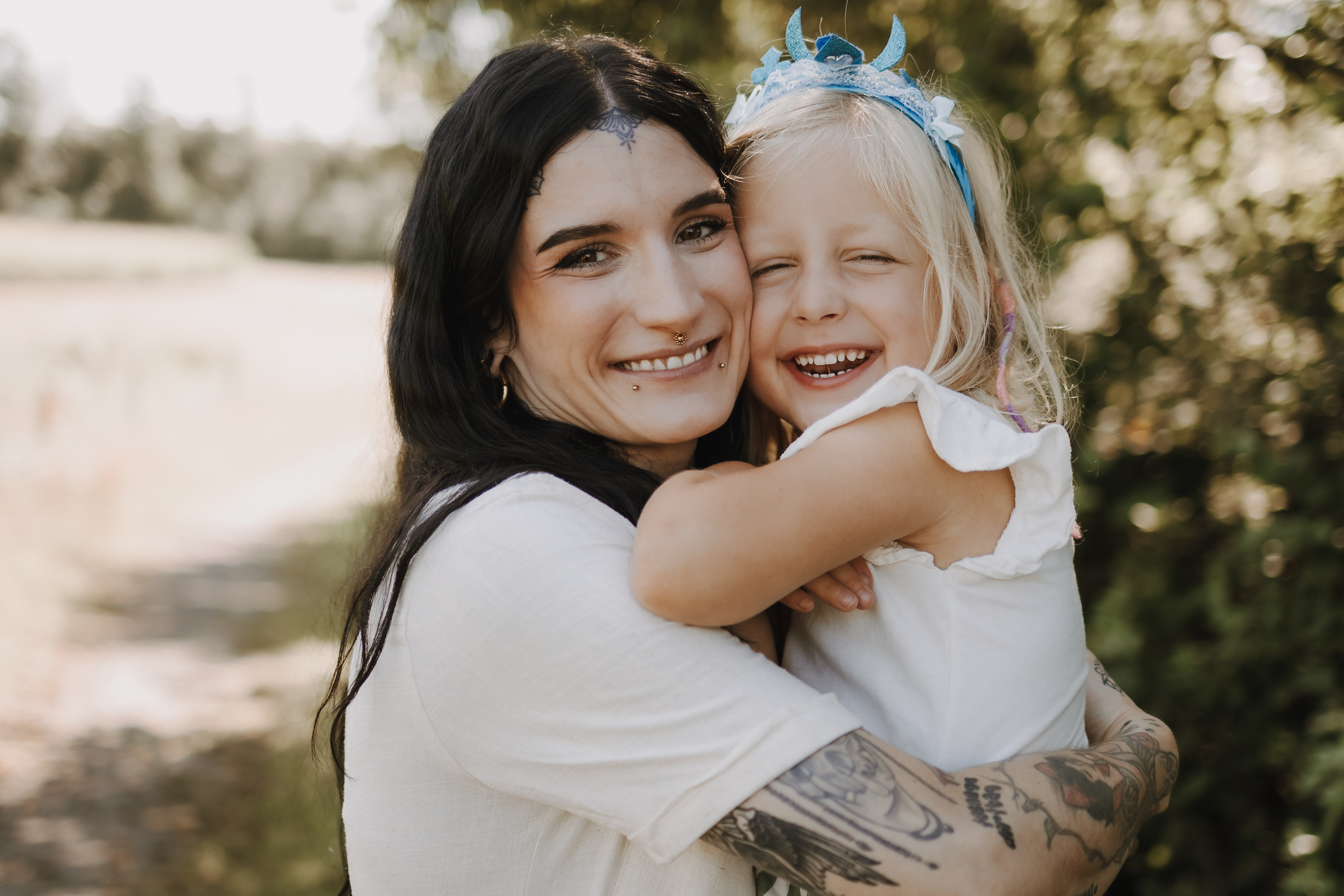 Eine Person mit langen dunklen Haaren und einem tätowierten Arm lächelt, während sie ein junges Mädchen mit blonden Haaren und einem blauen Stirnband umarmt. Sie sind an einem sonnigen Tag draußen mit Grün im Hintergrund und machen perfekte Familienfotos. Beide tragen weiße Oberteile und wirken glücklich und entspannt.