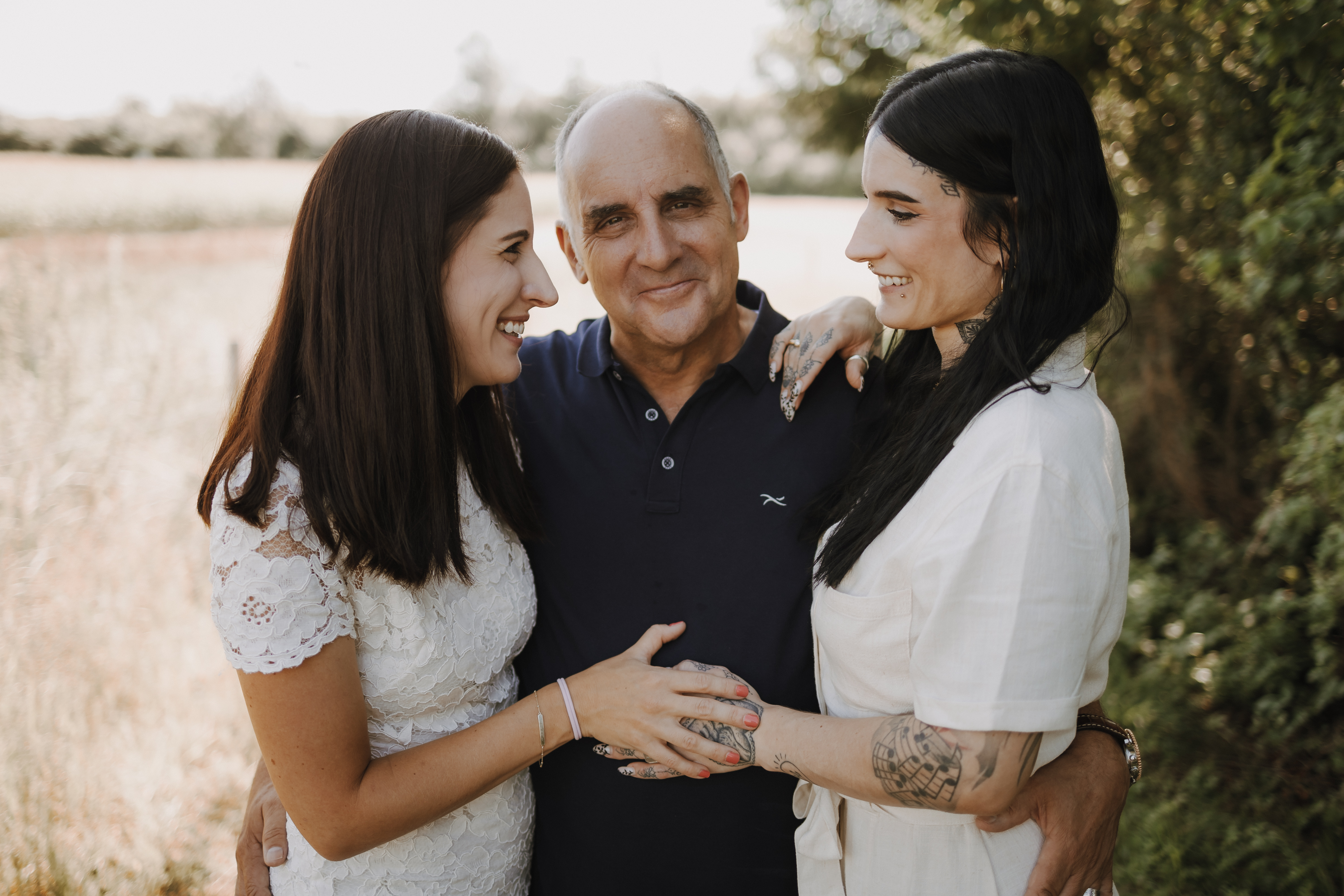 Ein kahlköpfiger Mann steht mit zwei Frauen, eine auf jeder Seite von ihm, im Freien während eines fröhlichen Familienshootings. Sie lächeln alle und wirken glücklich. Die Frau links trägt ein weißes Spitzenkleid, die Frau rechts hat Tattoos und trägt eine weiße Bluse. Im Hintergrund sind Bäume und Felder zu sehen.
