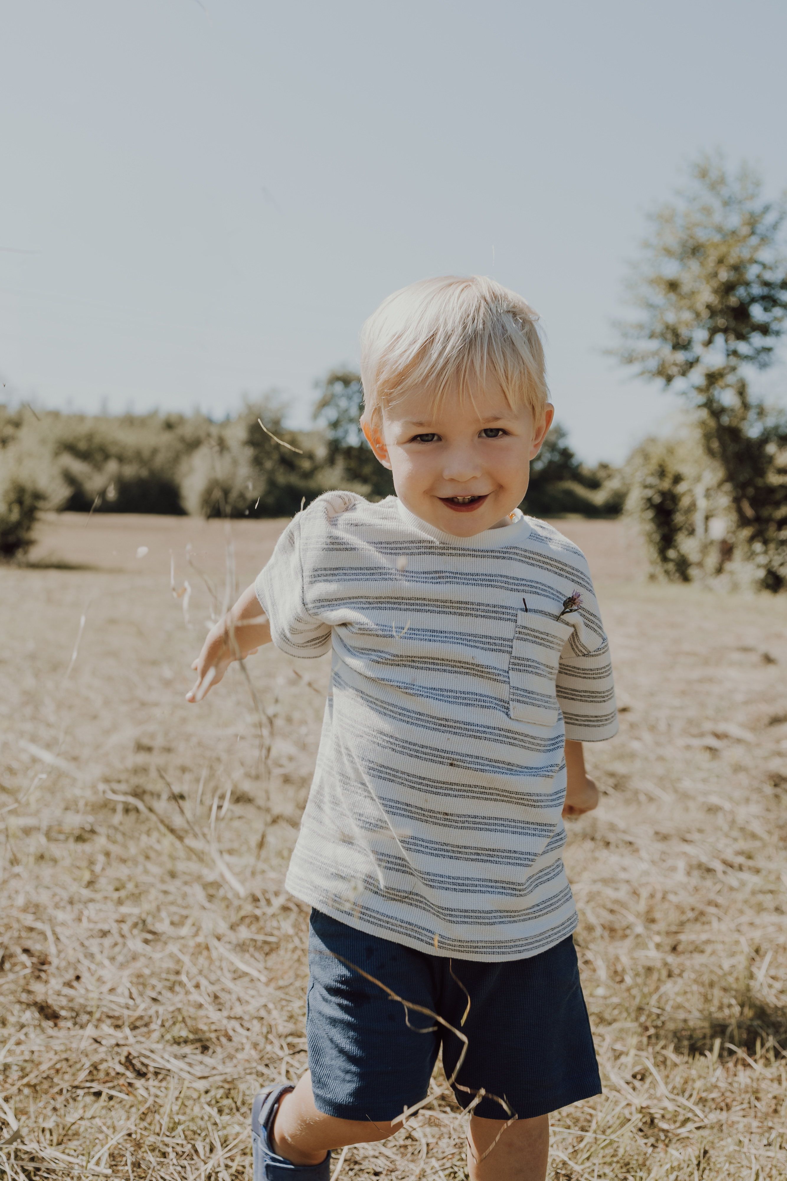 Ein kleiner Junge mit blonden Haaren läuft fröhlich lächelnd über eine Wiese. Er trägt ein gestreiftes Kurzarmhemd und dunkelblaue Shorts. Im Hintergrund sind ein klarer Himmel und Bäume in der Ferne zu sehen, perfekt für ein Fotoshooting in der Natur.