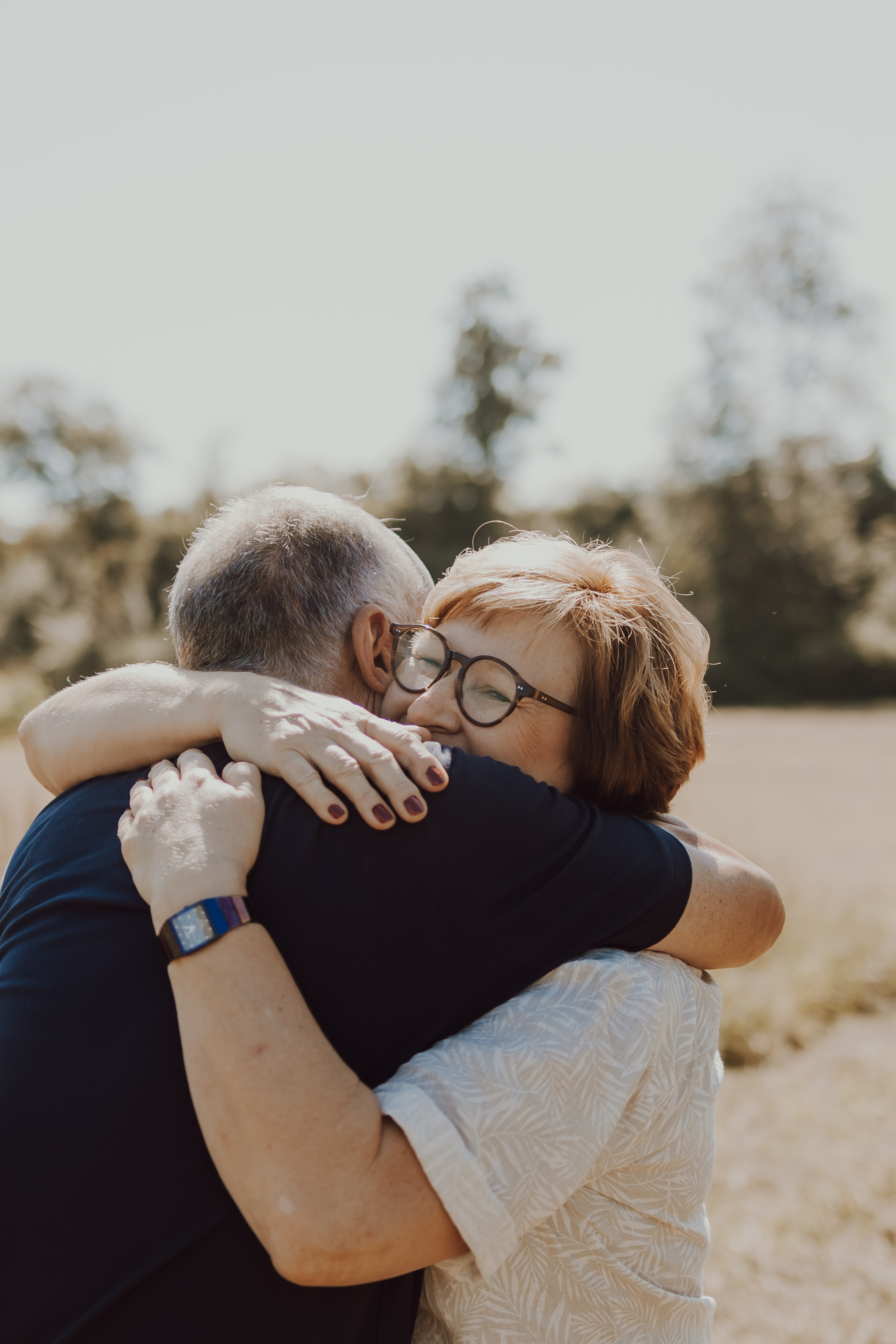 Ein älteres Paar umarmt sich freudig während eines Fotoshootings in der Natur. Die Frau, die eine Brille und ein weißes Hemd trägt, lächelt, während sie den Mann umarmt, der ein dunkles Hemd und eine Uhr trägt. Der Hintergrund ist ein sonnenbeschienenes Feld mit Bäumen, die in der Ferne verschwommen sind und ihren glücklichen Moment wunderschön einfangen.