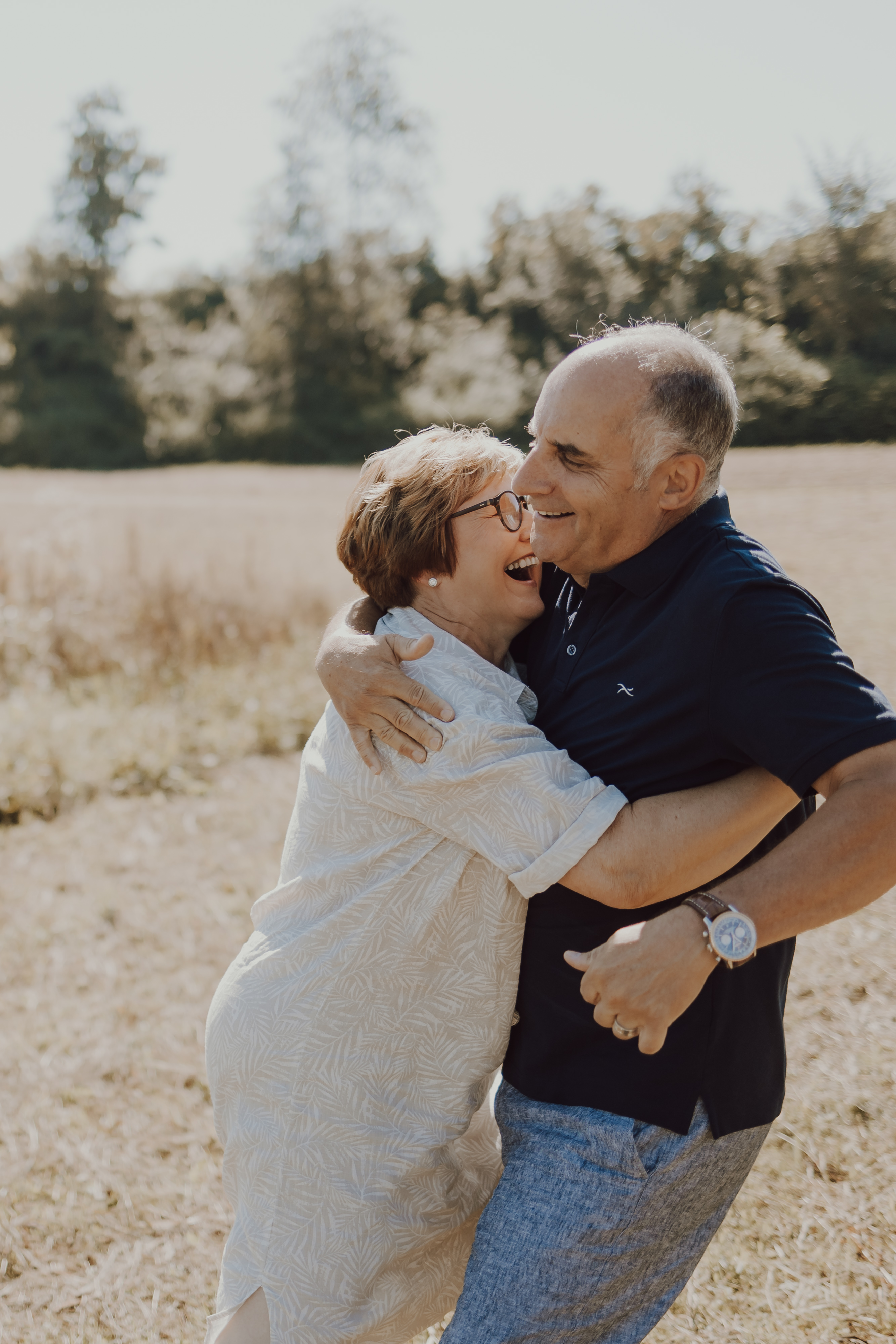 Ein fröhliches älteres Paar, eine Frau und ein Mann, umarmen sich und lachen gemeinsam im Freien während eines Fotoshootings. Sie stehen auf einer Wiese mit Bäumen im Hintergrund. Die Frau trägt eine Brille und ein helles Outfit; der Mann trägt ein dunkles Hemd und hat kurzes graues Haar. Dieser wunderbare Moment wurde von einer talentierten Fotografin in Baden festgehalten.