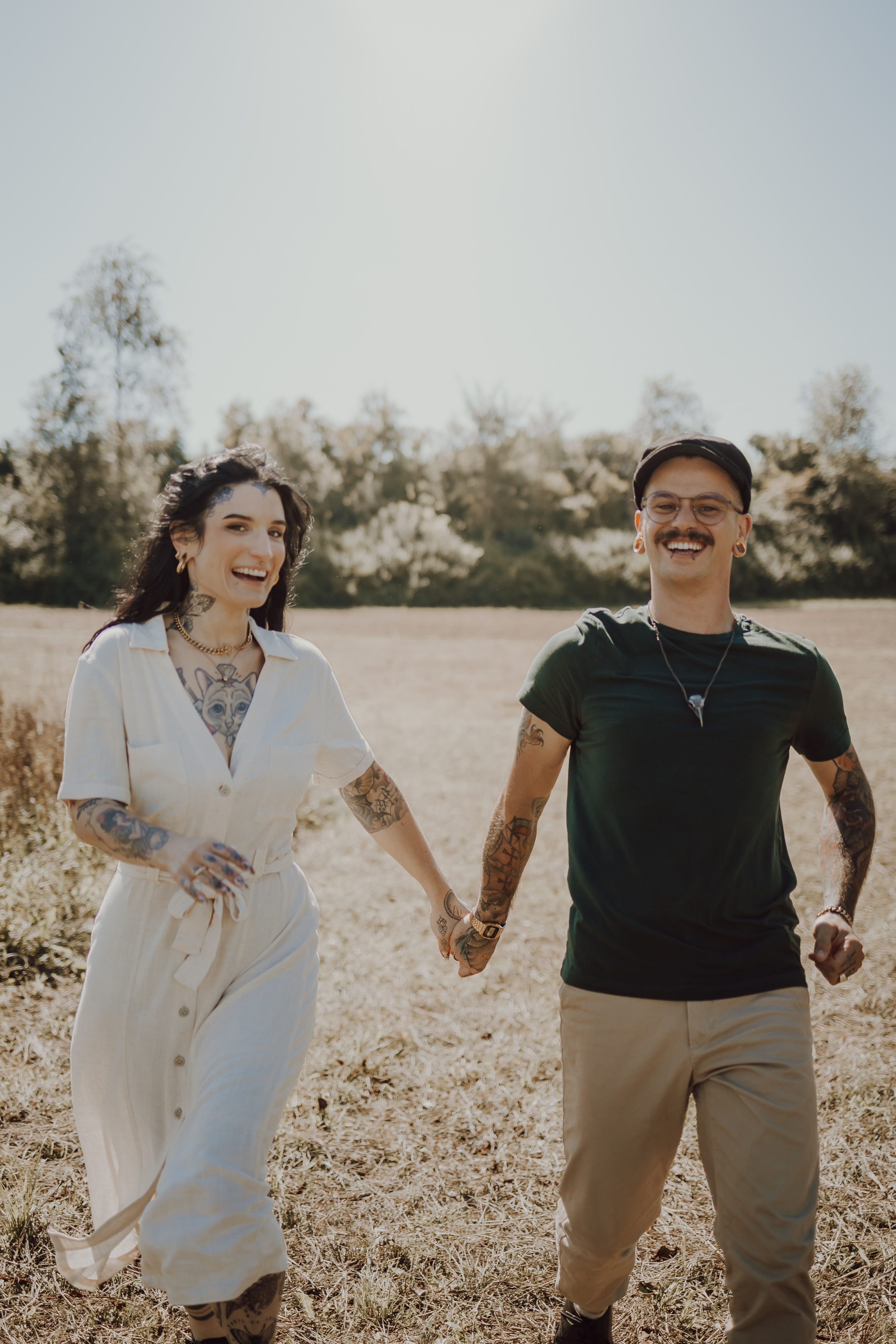 Eine Frau in einem weißen Kleid und ein Mann in einem grünen T-Shirt und einer Kappe, beide mit Tattoos, gehen Hand in Hand durch ein sonniges Feld, lächeln und sehen glücklich aus. Im Hintergrund sind Bäume unter einem klaren blauen Himmel zu sehen. Diese wunderschöne Szene wurde während eines Fotoshootings in der Natur mit Fotografin in Baden aufgenommen.