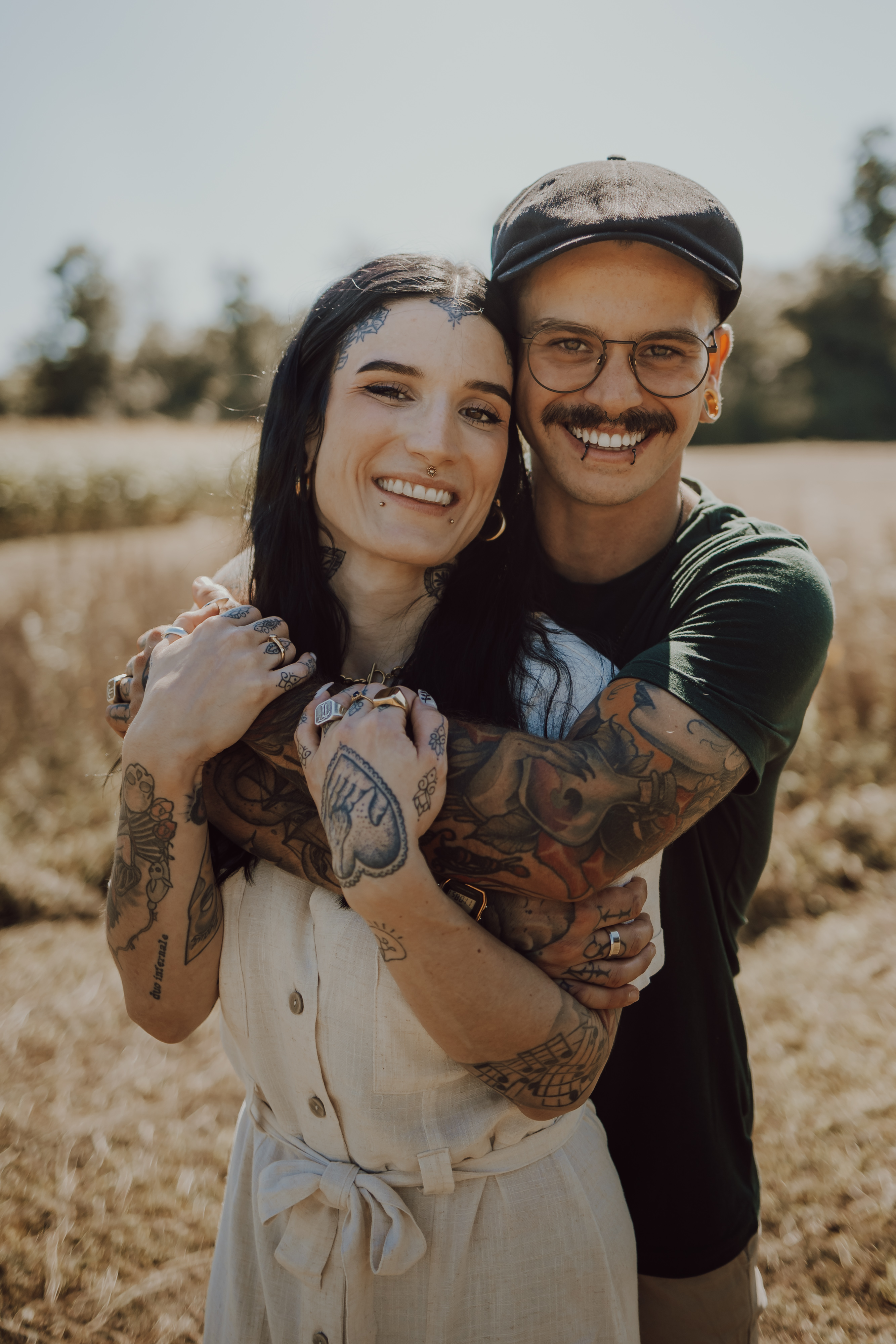 Ein lächelndes Paar steht auf einem Feld mit hohem Gras und Bäumen im Hintergrund, wunderschön eingefangen von einer Fotografin in Baden. Die Frau trägt ein helles Kleid, hat dunkle Haare und Tattoos auf den Armen. Der Mann, der eine Brille, eine Mütze und ein grünes Hemd trägt, umarmt sie von hinten und zeigt ebenfalls seine Tattoos.