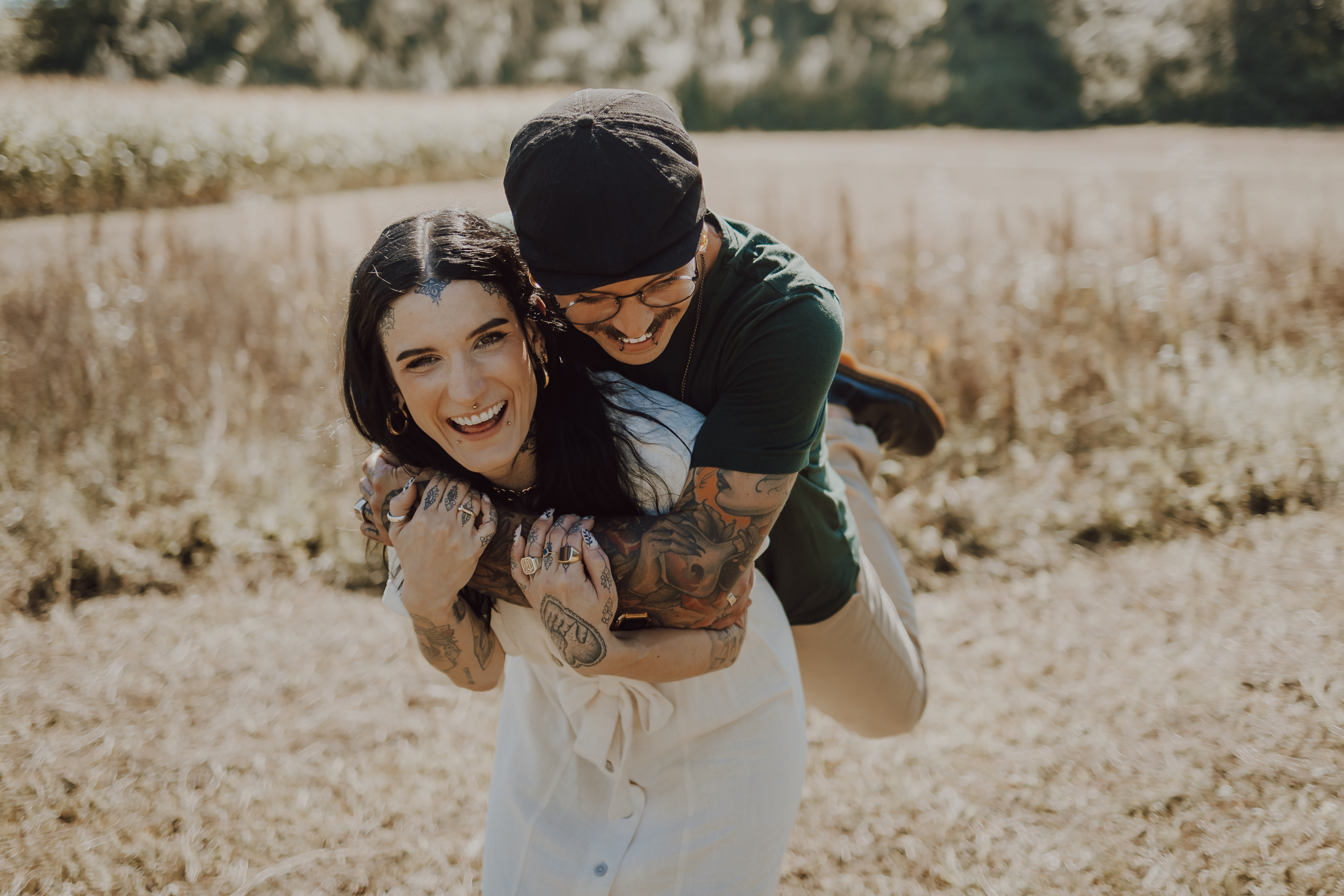 Zwei Menschen genießen einen Moment im Freien, wobei einer den anderen Huckepack trägt und beide breit lächeln, als ob sie auf einem fröhlichen Familienfoto wären. Die Person, die getragen wird, trägt ein weißes Kleid, während die Person, die sie trägt, ein grünes Hemd, beige Hosen und eine schwarze Mütze trägt und sichtbare Tattoos hat.