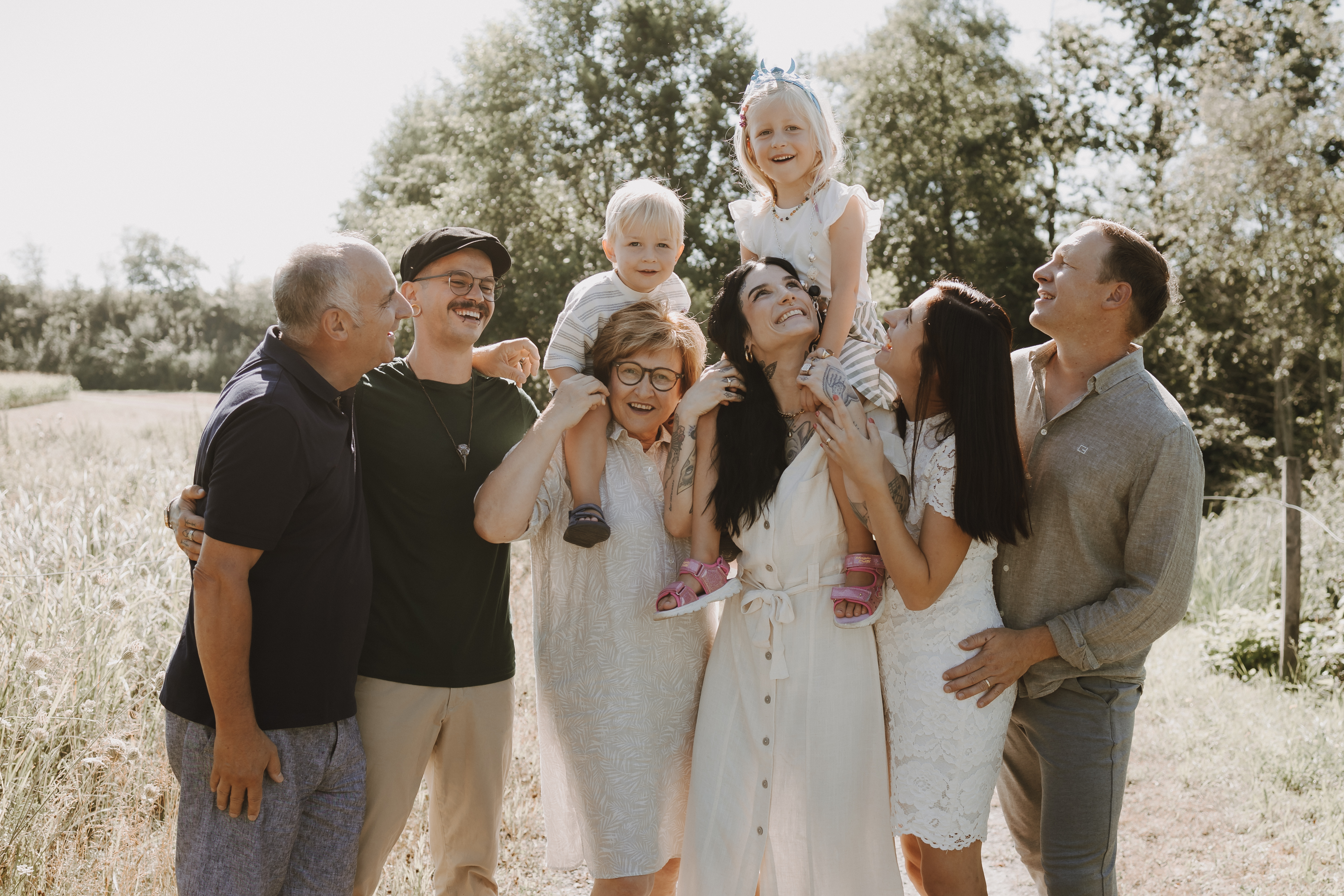 Eine fröhliche Mehrgenerationenfamilie steht bei einem Fotoshooting in der Natur auf einer sonnigen Wiese zusammen. Zwei Kinder werden fröhlich von Erwachsenen gehalten, während alle lächeln und lachen. Im Hintergrund sind Bäume und ein blauer Himmel zu sehen, die die fröhliche Atmosphäre im Freien verstärken.