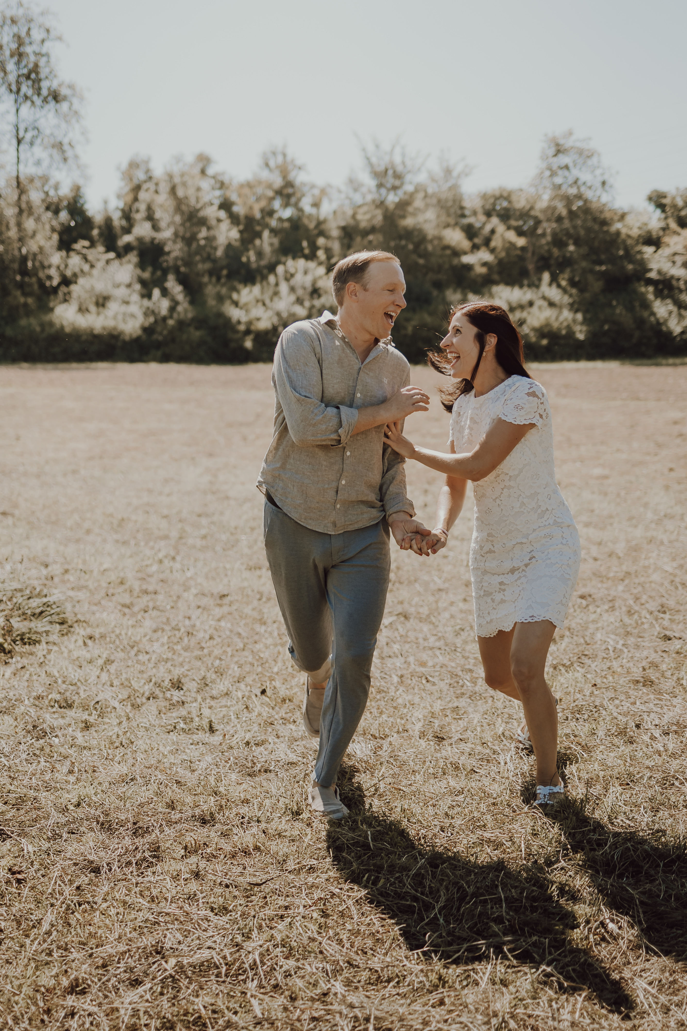 Ein Paar hält Händchen und läuft fröhlich über eine Wiese während seines Familienshootings. Der Mann trägt ein graues Outfit und die Frau ein kurzes weißes Spitzenkleid. Sie lächeln und scheinen einen hellen, sonnigen Tag mit Bäumen im Hintergrund und klarem Himmel zu genießen.