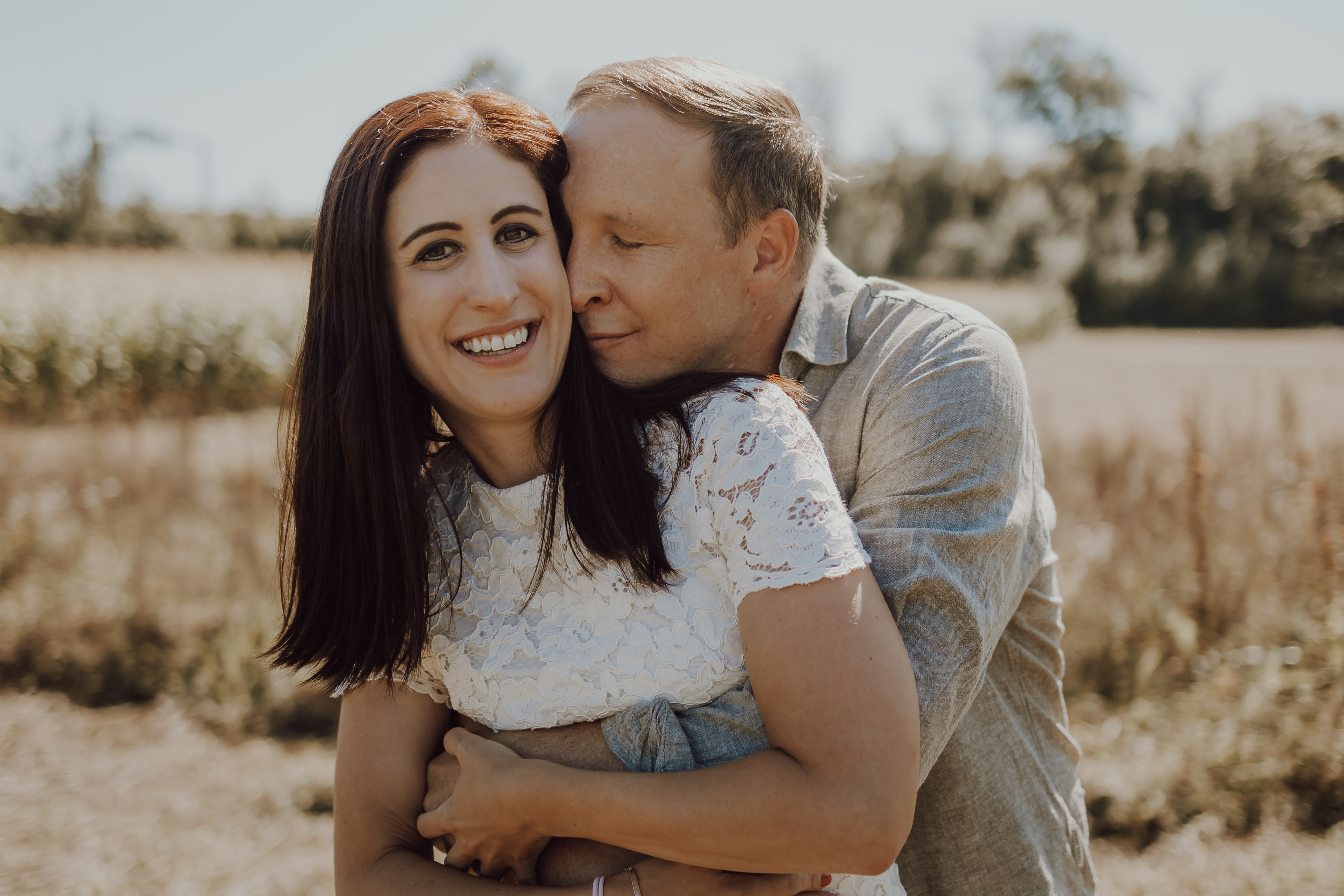Eine Frau mit langen braunen Haaren, die ein weißes Spitzenkleid trägt, lächelt, während sie von hinten von einem Mann mit kurzen Haaren und einem grauen Hemd umarmt wird. Während ihres Fotoshootings in der Natur stehen sie auf einem sonnenbeschienenen Feld mit hohem Gras und Bäumen in der Ferne.