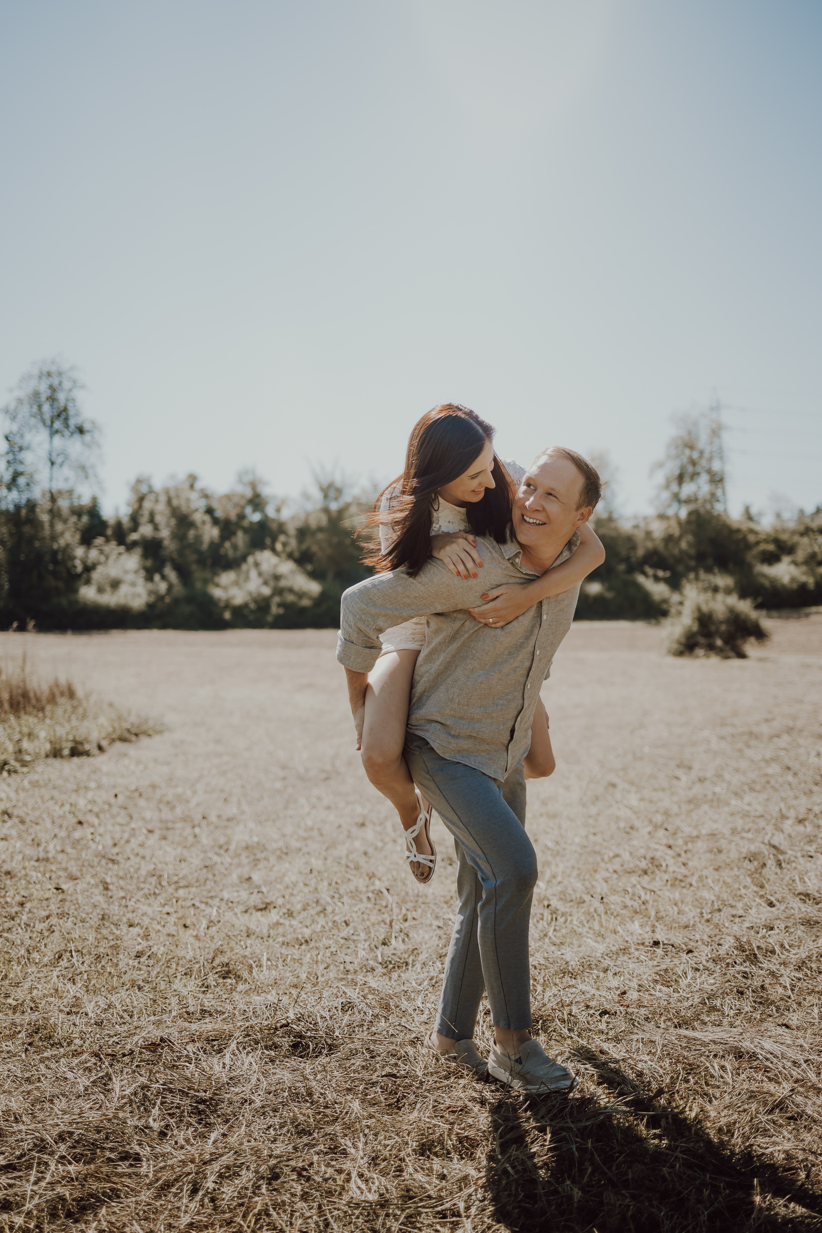 Inmitten eines sonnenbeschienenen offenen Feldes reitet eine Person freudig Huckepack auf einer anderen, beide lächeln strahlend. Bäume und Sträucher säumen den Hintergrund unter einem klaren blauen Himmel. Es ist die perfekte Kulisse für Familienfotos während eines Fotoshootings in der Natur mit Ihrer Fotografin in Baden.