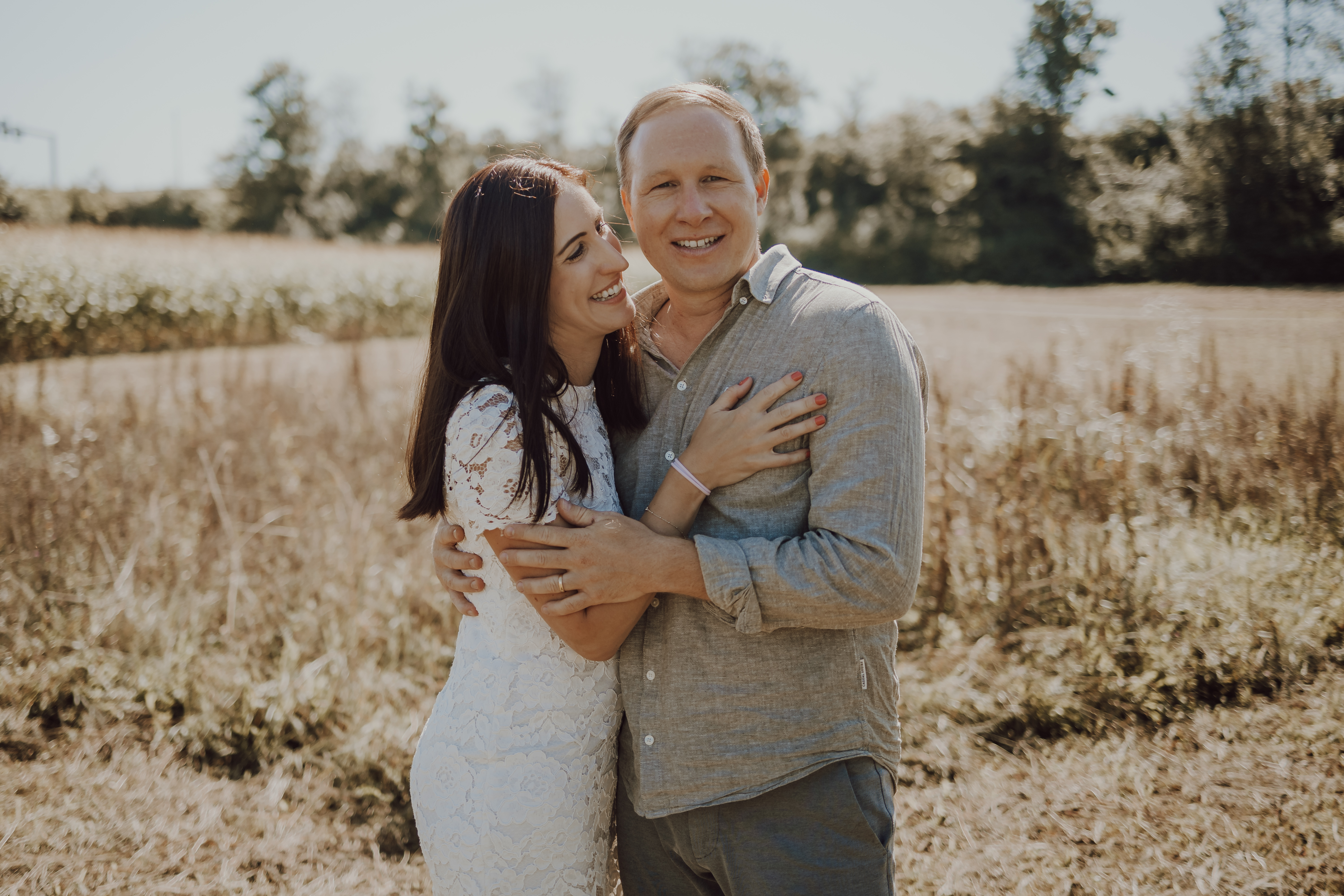 Ein Paar steht bei einem Fotoshooting in der Natur eng aneinander gekuschelt auf einer sonnenbeschienenen Wiese. Die Frau im weißen Spitzenkleid umarmt den Mann, der ein hellgraues Hemd trägt. Beide lächeln sich herzlich an. Im Hintergrund sind hohe Gräser und Bäume zu sehen, die eine malerische Landschaft suggerieren.