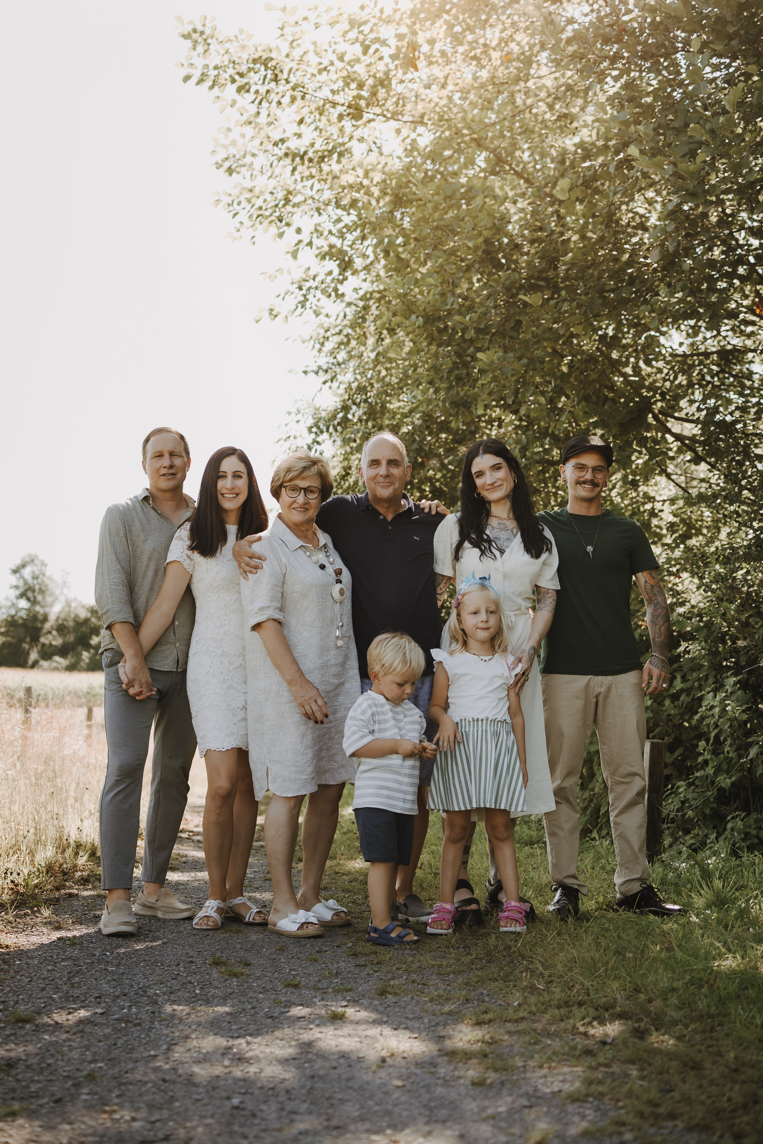 Eine siebenköpfige Familie steht an einem sonnigen Tag im Freien und posiert für ein Familienshooting. Die Gruppe besteht aus drei Erwachsenen, einer älteren Frau, einer jungen brünetten Frau, die ein blondes Mädchen an der Hand hält, und einem Mann, der hinter einem kleinen Jungen steht. Bei diesem wunderschönen Fotoshooting in der Natur sind Bäume und Grasfelder im Hintergrund zu sehen.