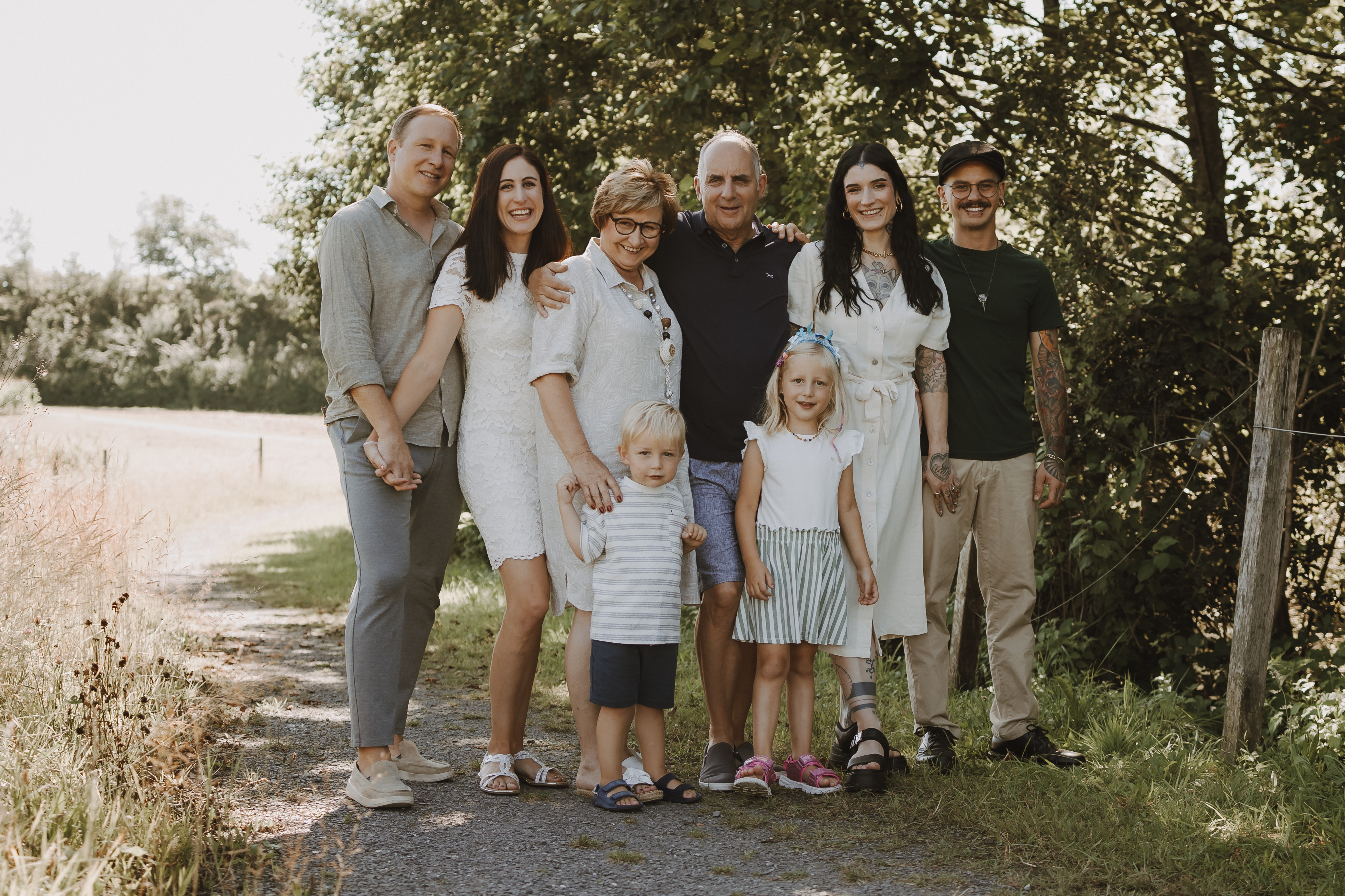Eine Gruppe von acht Personen, bestehend aus Männern, Frauen und Kindern, posiert an einem sonnigen Tag im Freien. Sie stehen auf einem Kiesweg mit hohem Gras und Bäumen im Hintergrund. Einige halten Händchen und alle lächeln bei diesem fröhlichen Familienshooting.