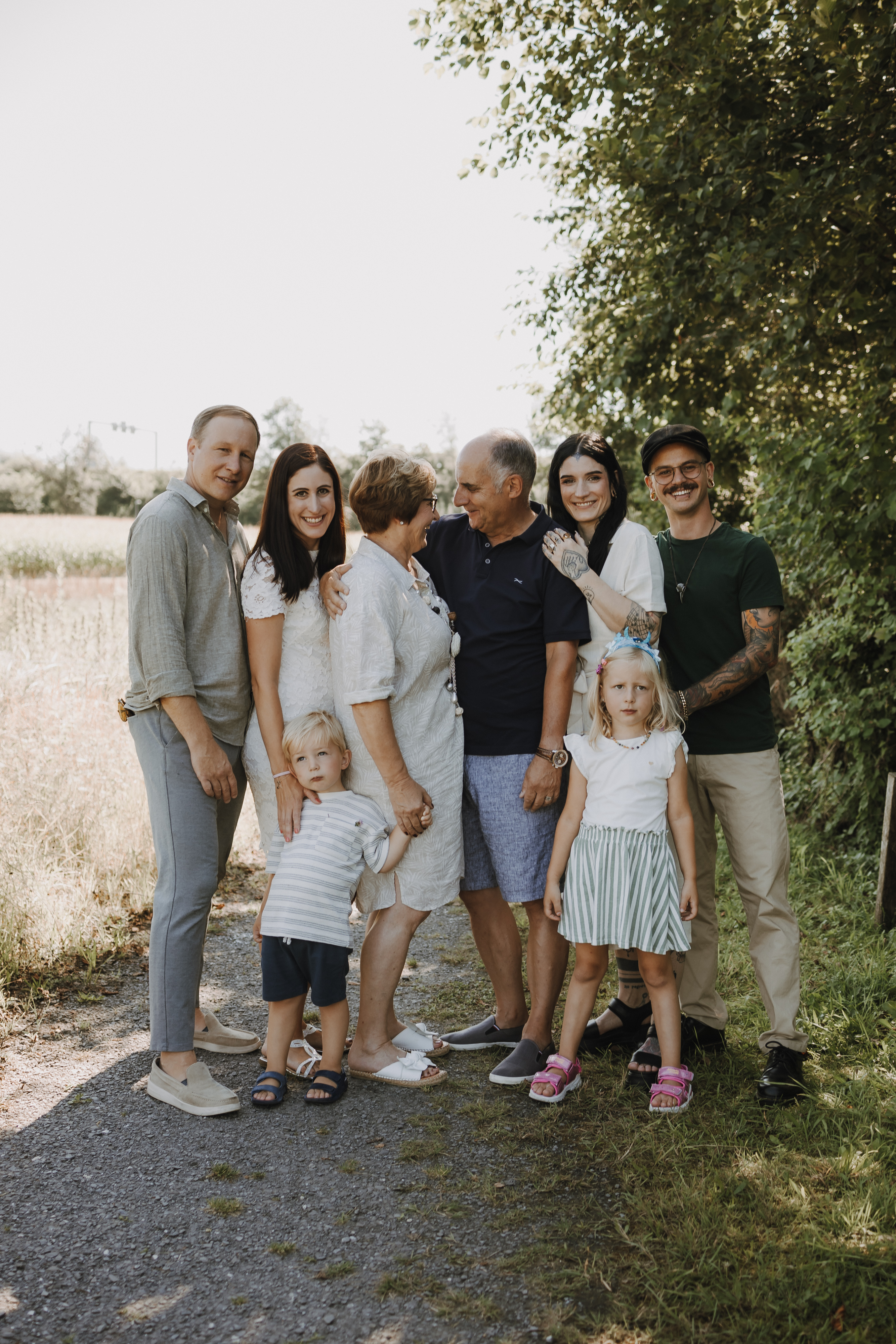 Eine achtköpfige Familie posiert an einem sonnigen Tag im Freien. Die Gruppe besteht aus Erwachsenen, zwei kleinen Kindern und älteren Personen. Sie stehen auf einem von Grün umgebenen Kiesweg, lächeln und sehen glücklich aus – perfekt für Familienfotos während eines Fotoshootings in der Natur.
