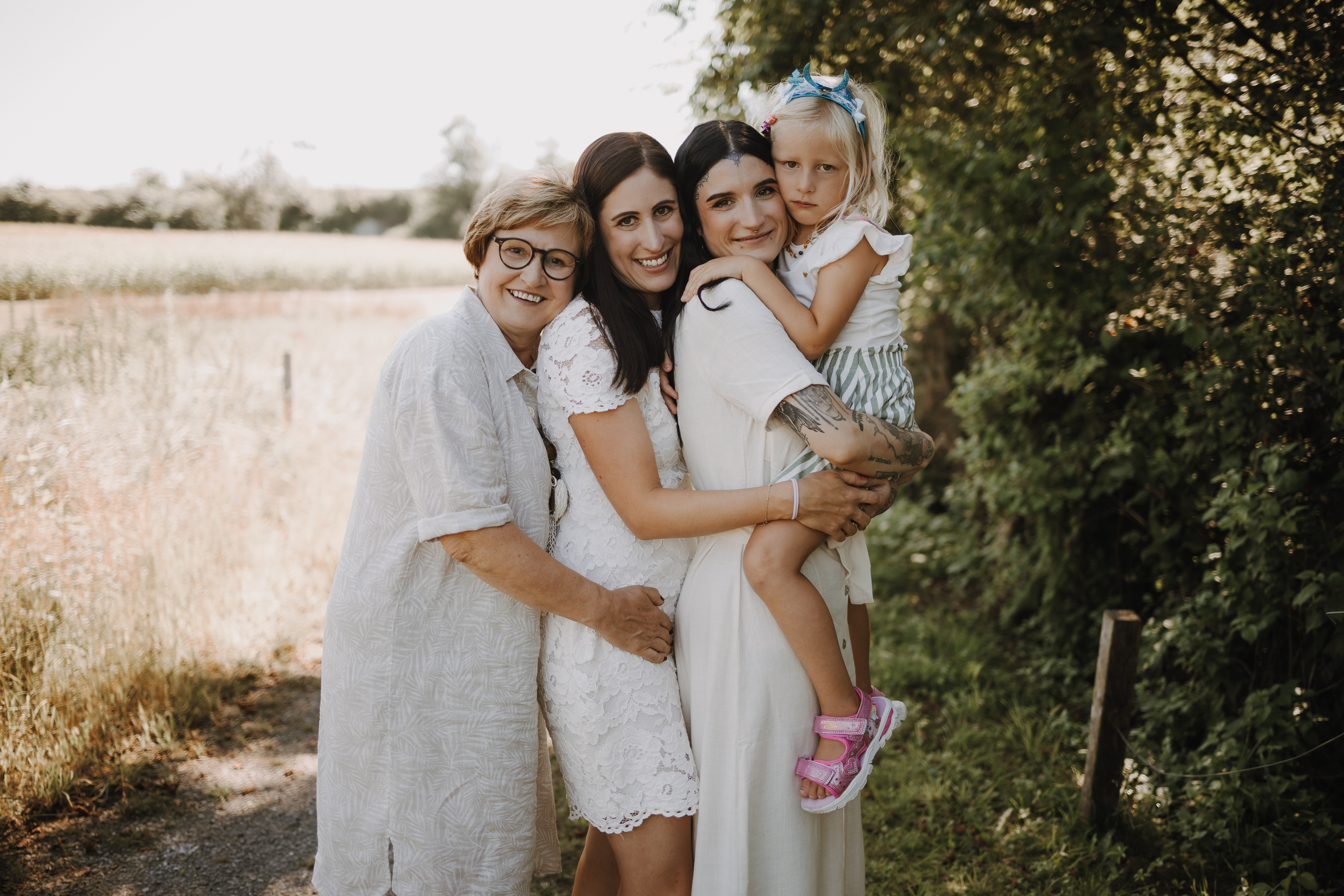 Drei Frauen und ein junges Mädchen stehen bei einem entzückenden Fotoshooting in der Natur dicht beieinander und lächeln in einer Außenkulisse mit Bäumen und Grün. Eine Frau hält das Kind, das ein gestreiftes Kleid und rosa Schuhe trägt. Die Frauen tragen helle Kleidung. Der Hintergrund ist sonnenbeschienen.