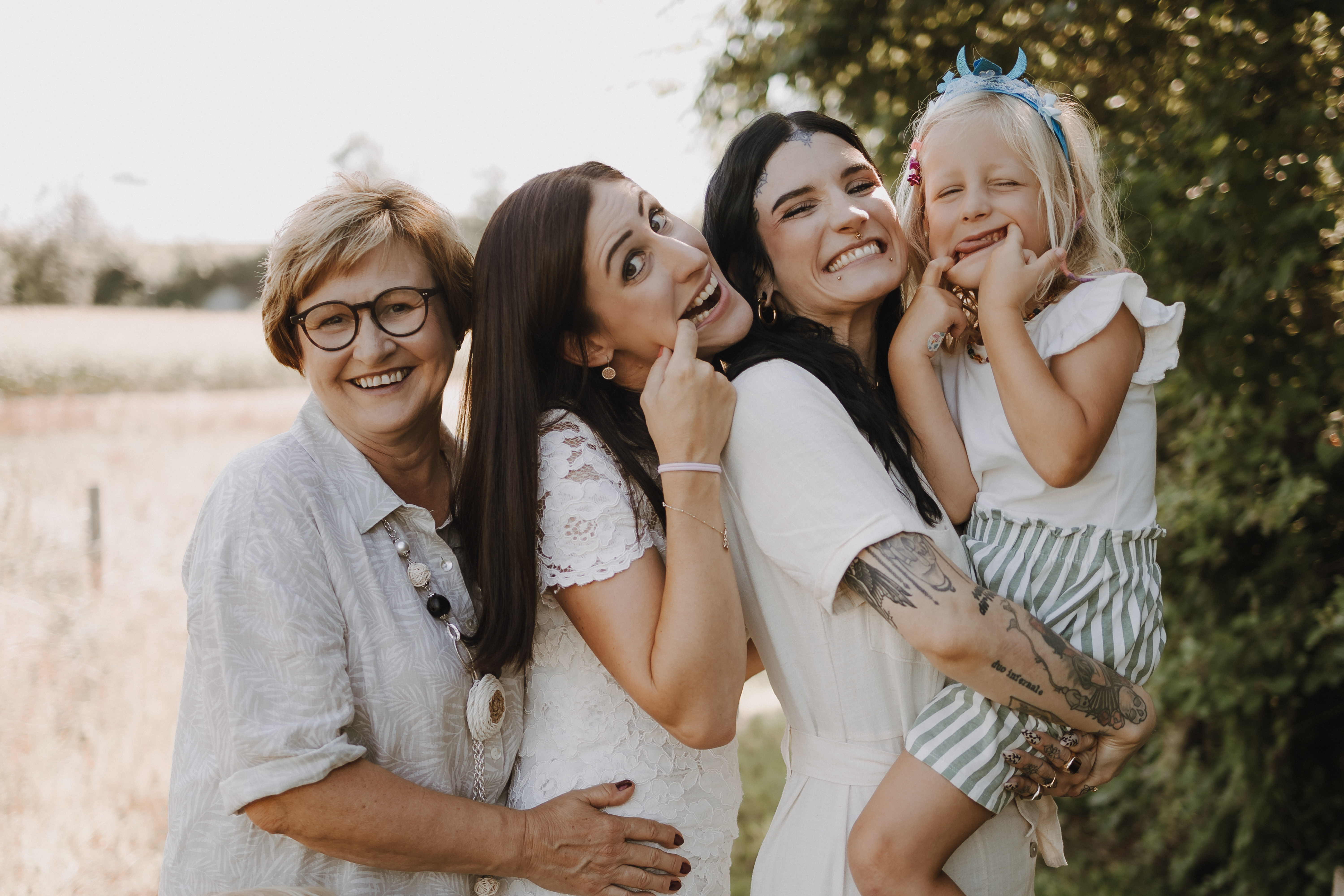 Eine Gruppe von vier Personen steht im Freien und lächelt während eines Familienshootings in die Kamera. Eine ältere Frau mit Brille, zwei junge Frauen und ein junges Mädchen mit einem blauen Stirnband. Das Mädchen wird von einer der Frauen hochgehalten und alle wirken glücklich. Im Hintergrund sind ein Feld und Bäume zu sehen.