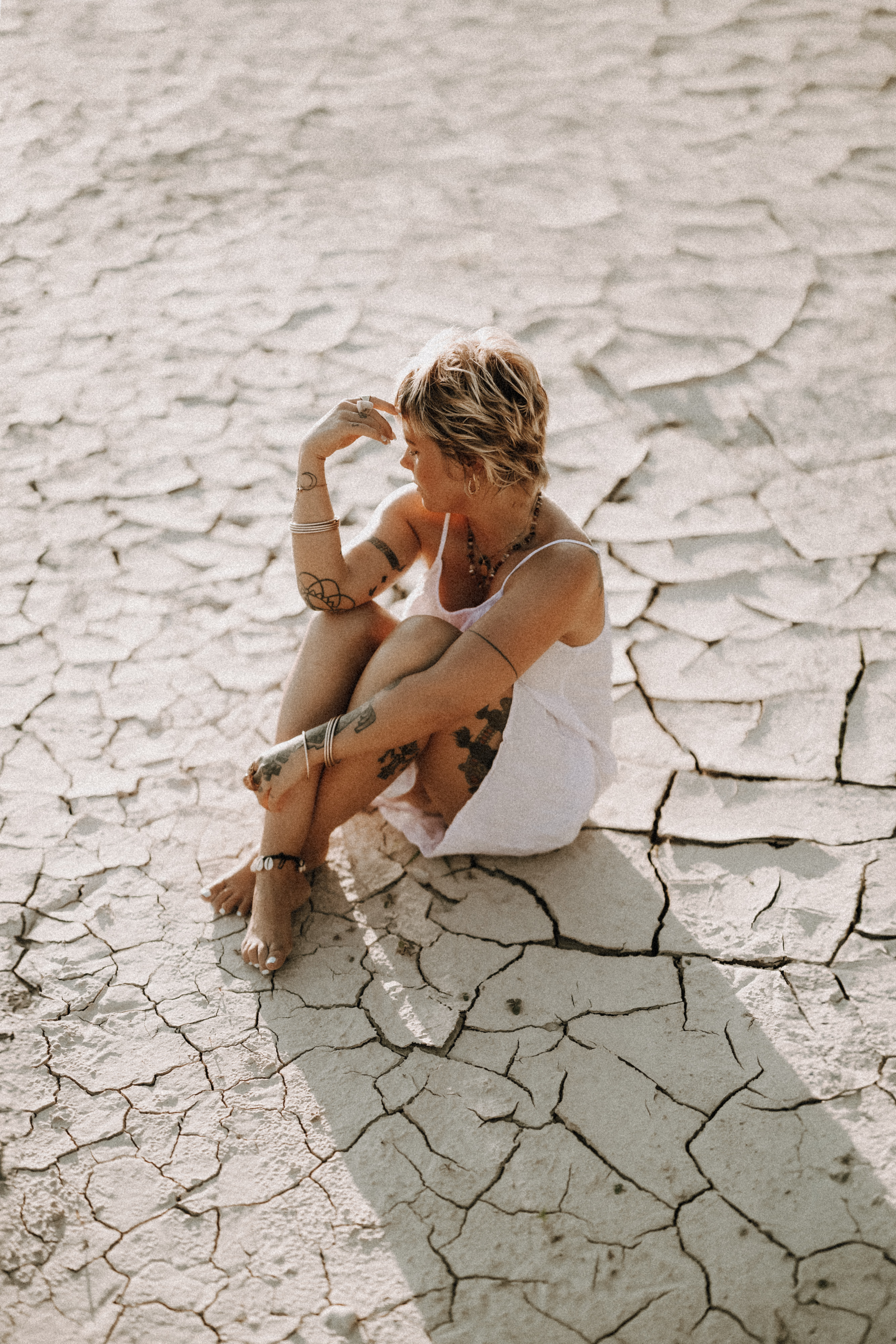 Eine Person mit kurzen Haaren, einem weißen Kleid und verschiedenen Tattoos sitzt in einer rissigen, trockenen Landschaft. Sie blickt zur Seite, eine Hand berührt ihr Gesicht und die andere ruht auf ihrem Knie. Bei diesem atemberaubenden Fotoshooting in Baden wirft die tief stehende Sonne Schatten auf den Boden.