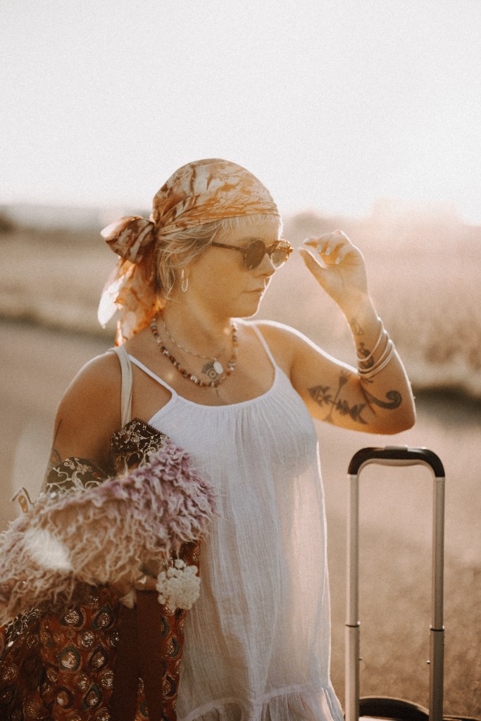 Eine Person mit Sonnenbrille und gemustertem Kopftuch schiebt einen Gepäckwagen eine sonnenbeschienene Straße entlang. Sie trägt ein weißes ärmelloses Kleid, hat Tattoos auf dem linken Arm und trägt eine bunte Tasche mit Quasten. Diese selbstbewusste Person strahlt Selbstbewusstsein aus, in einer Umgebung, die vor dem unscharfen Hintergrund im Freien die perfekte Porträtsituation sein könnte.