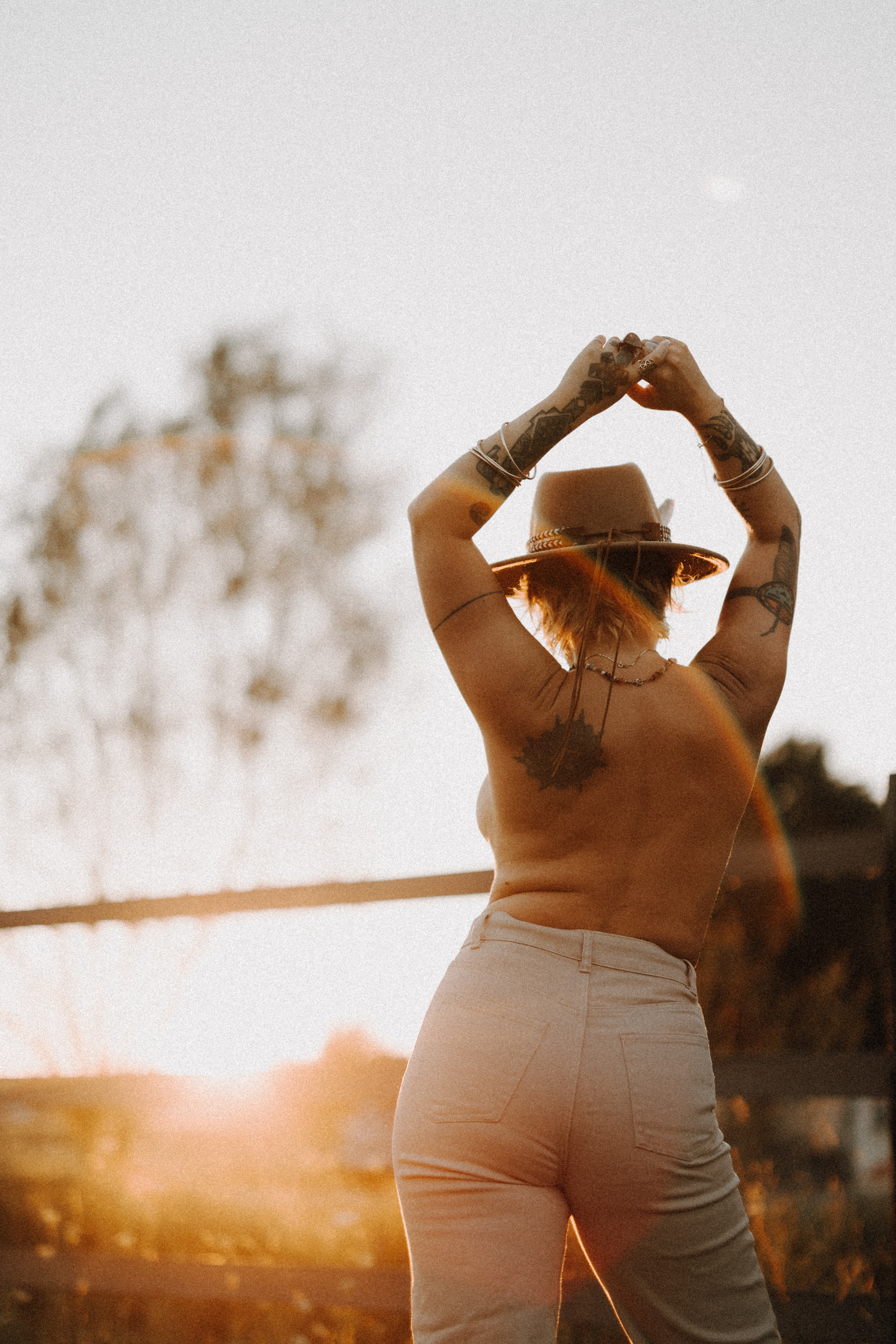 Eine Person mit Tattoos steht bei Sonnenuntergang im Freien, blickt von der Kamera weg, hat die Arme erhoben und trägt einen Hut. Das Sonnenlicht erzeugt einen warmen Schein um sie herum und im Hintergrund sind ein verschwommener Baum und ein Zaun zu sehen – es ist ein intimes Porträt, das die Essenz einer spontanen Fotoshooting-Session einfängt.