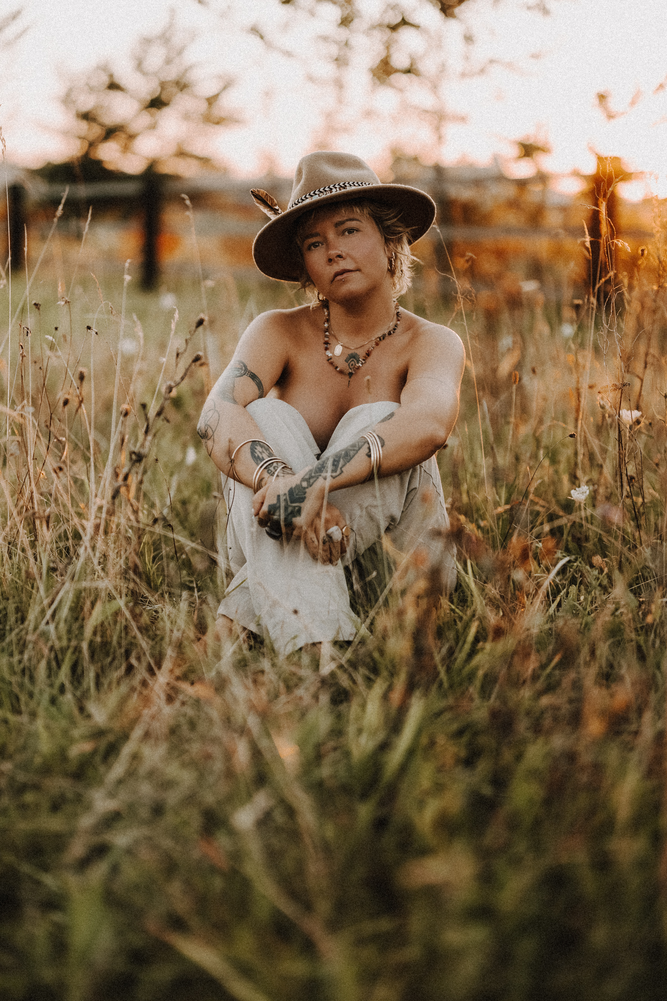 Eine Person mit kurzen Haaren, einem breitkrempigen Hut, Halsketten und Armbändern sitzt im Schneidersitz bei Sonnenuntergang auf einer Wiese. Die Person ist barfuß und hat Tattoos auf den Armen, umgeben von hohem Gras und einem unscharfen natürlichen Hintergrund – perfekt für ein inspirierendes Fotoshooting in Baden.