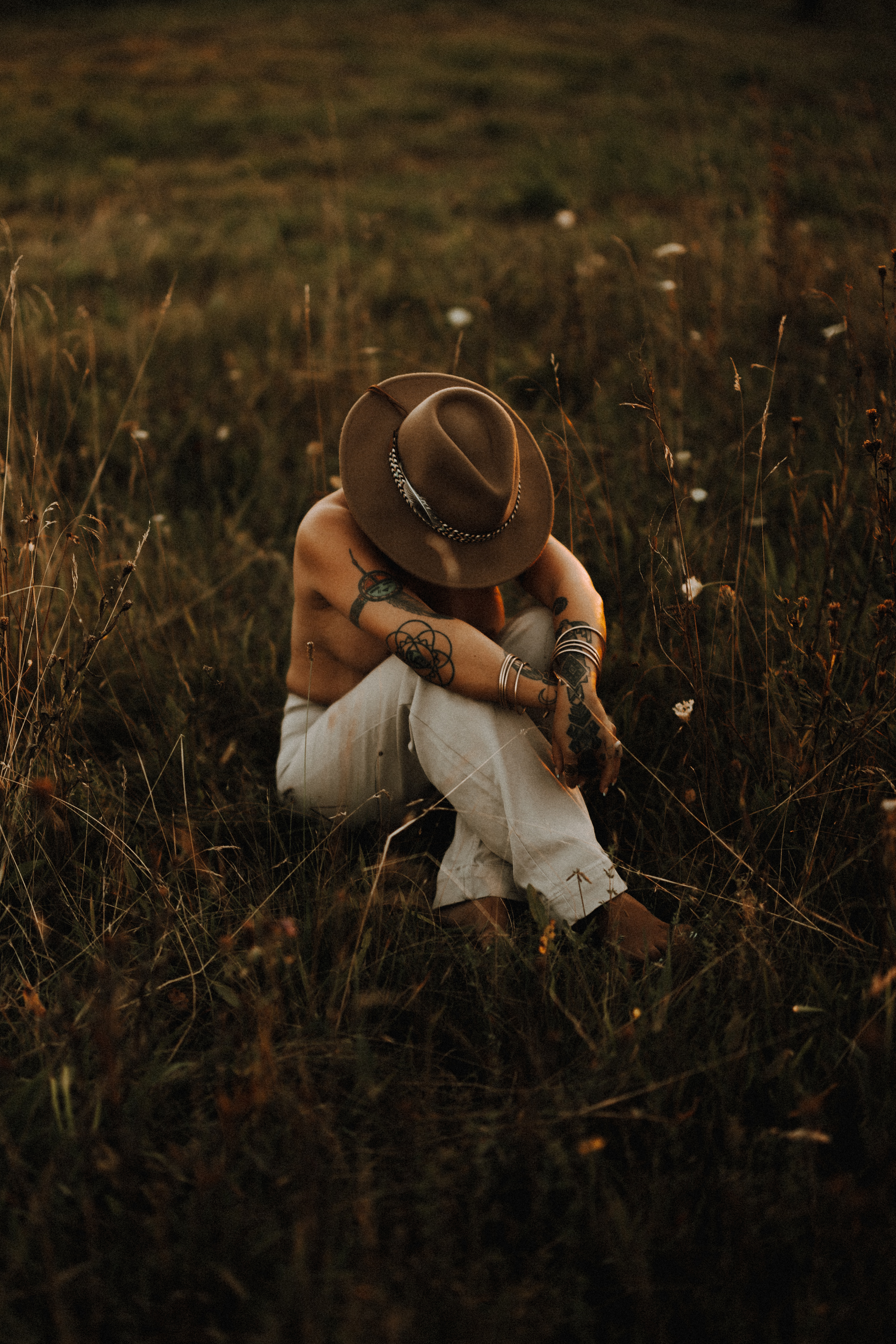Eine Person mit breitkrempigem Hut und weißer Hose sitzt während eines Fotoshootings in Baden in einem Feld mit hohem Gras und Wildblumen. Ihr Kopf ist gesenkt und ihre tätowierten, mit Armbändern geschmückten Arme ruhen in einer nachdenklichen Pose auf ihren Knien. Die untergehende Sonne wirft einen goldenen Schimmer auf die Szene.