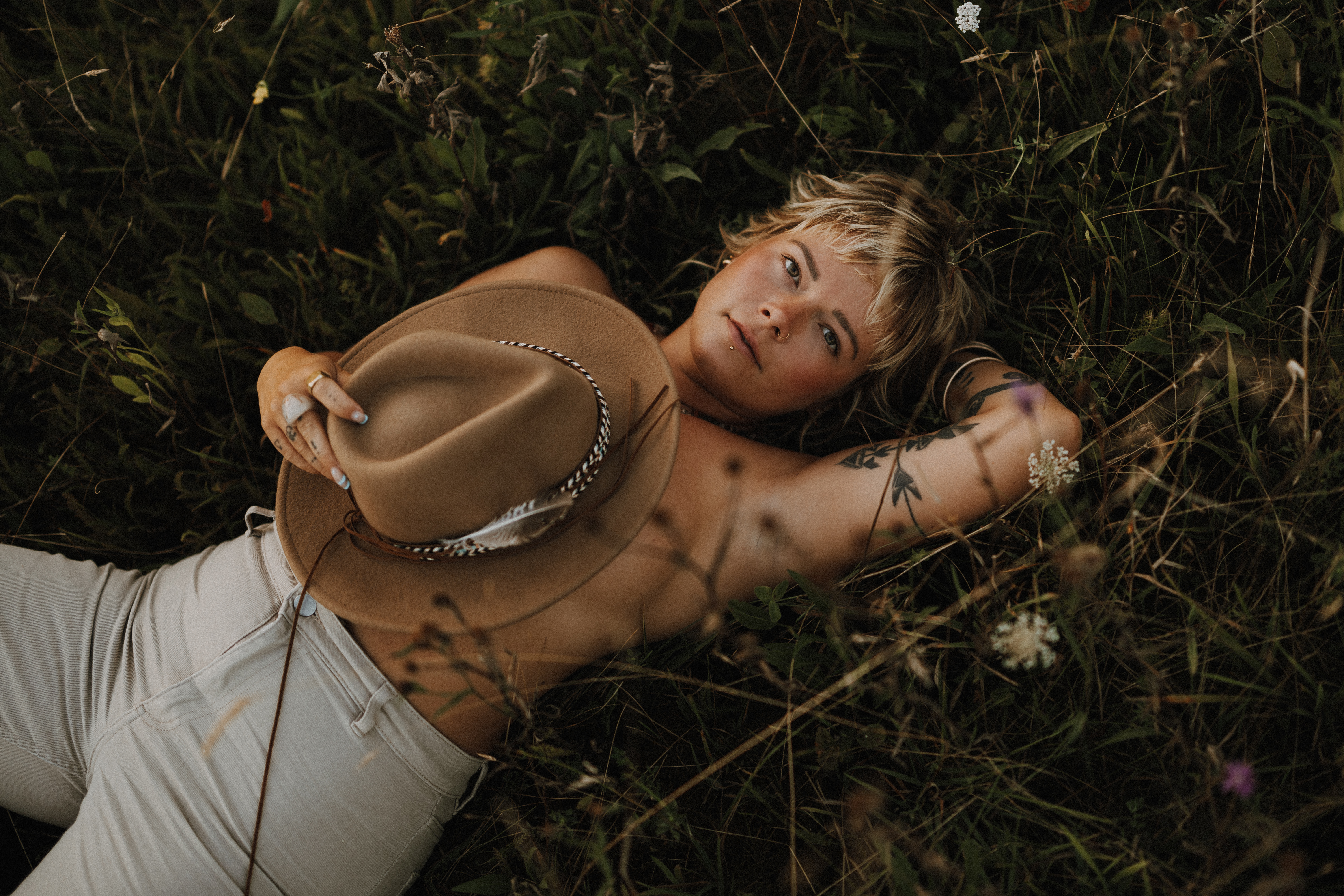 Eine Person mit kurzen blonden Haaren und Tattoos liegt auf einer Wiese, die teilweise von Grünpflanzen und Wildblumen bedeckt ist. Sie hält einen braunen Hut, der ihren Körper teilweise bedeckt, und blickt nach oben. Sie trägt weiße Hosen und ist mit Ringen und Armbändern geschmückt – perfekt für ein Fotoshooting in Baden mit einem erfahrenen Fotografen oder einer erfahrenen Fotografin.