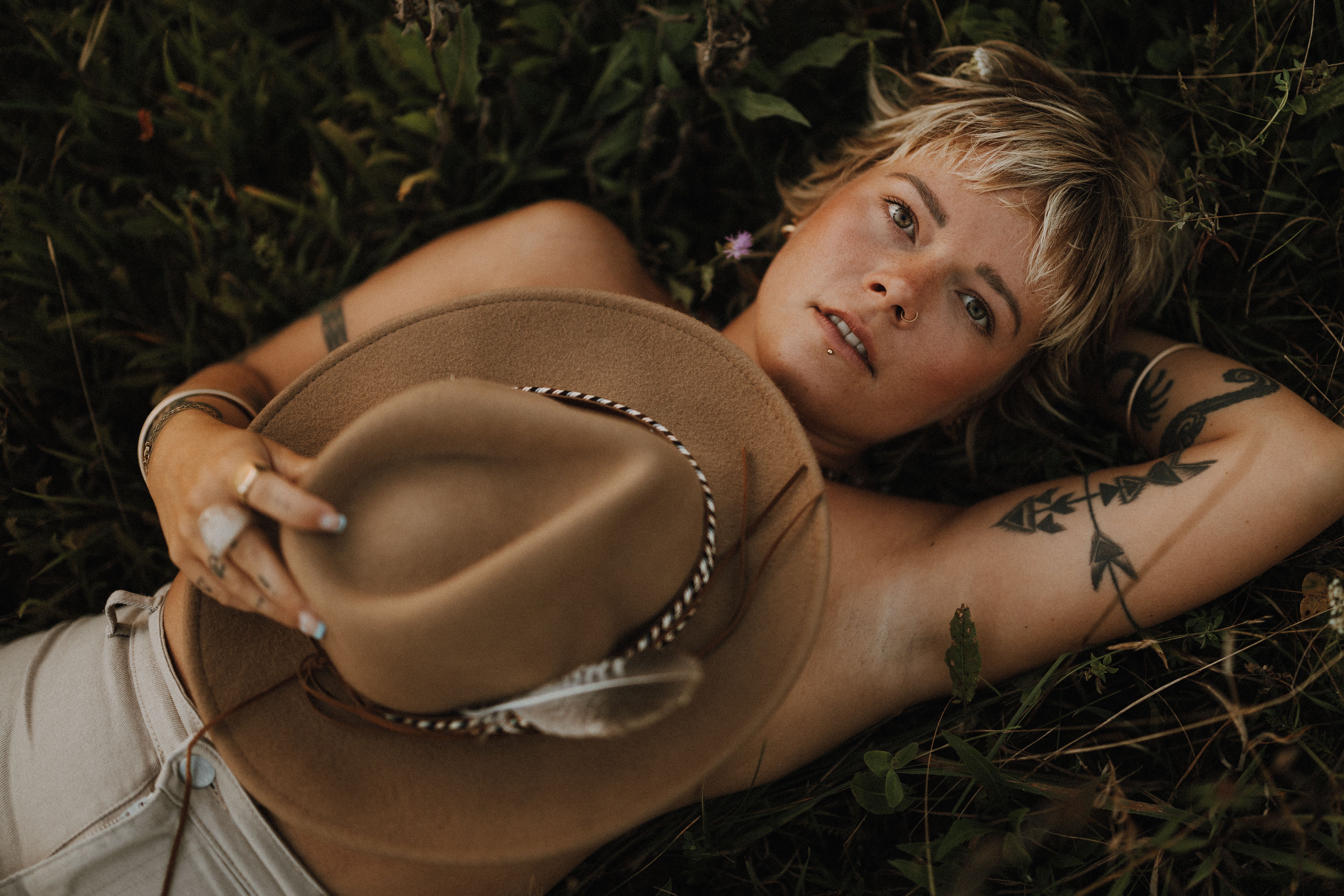 Eine Person mit kurzen blonden Haaren und Tattoos liegt auf einer Wiese und hält einen braunen Hut mit einer Feder in der Hand. Sie hat einen nachdenklichen Gesichtsausdruck und ist von Grün und Wildblumen umgeben. Sie fängt die Essenz eines natürlichen Fotoshooting-Moments ein.