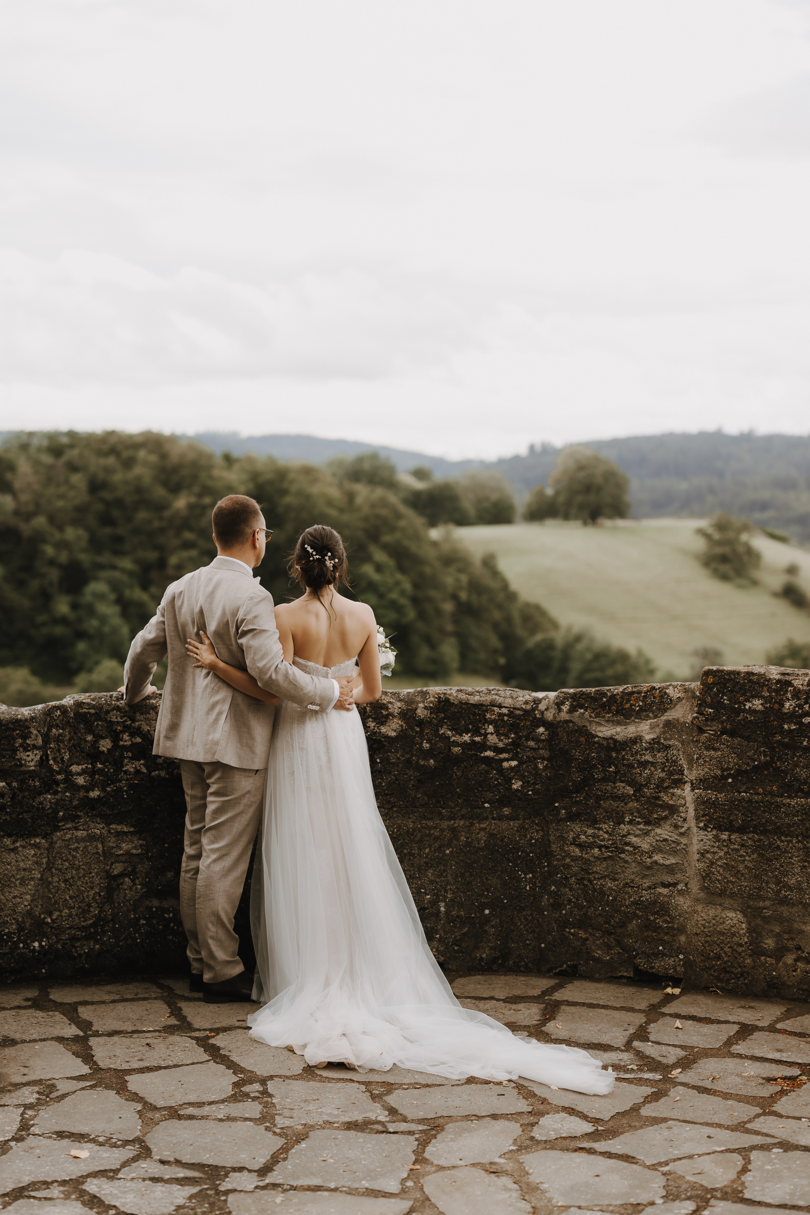 Ein Paar steht zusammen auf einer Steinterrasse mit Blick auf eine malerische Landschaft im Aargau. Der Mann, der einen hellgrauen Anzug trägt, hat seinen Arm um die Taille der Frau gelegt, die ein fließendes weißes Hochzeitskleid aus der Hochzeitsfotografie-Kollektion von Tasha Ender Photography trägt. Sie blicken auf sanfte grüne Hügel und Bäume unter einem bewölkten Himmel.