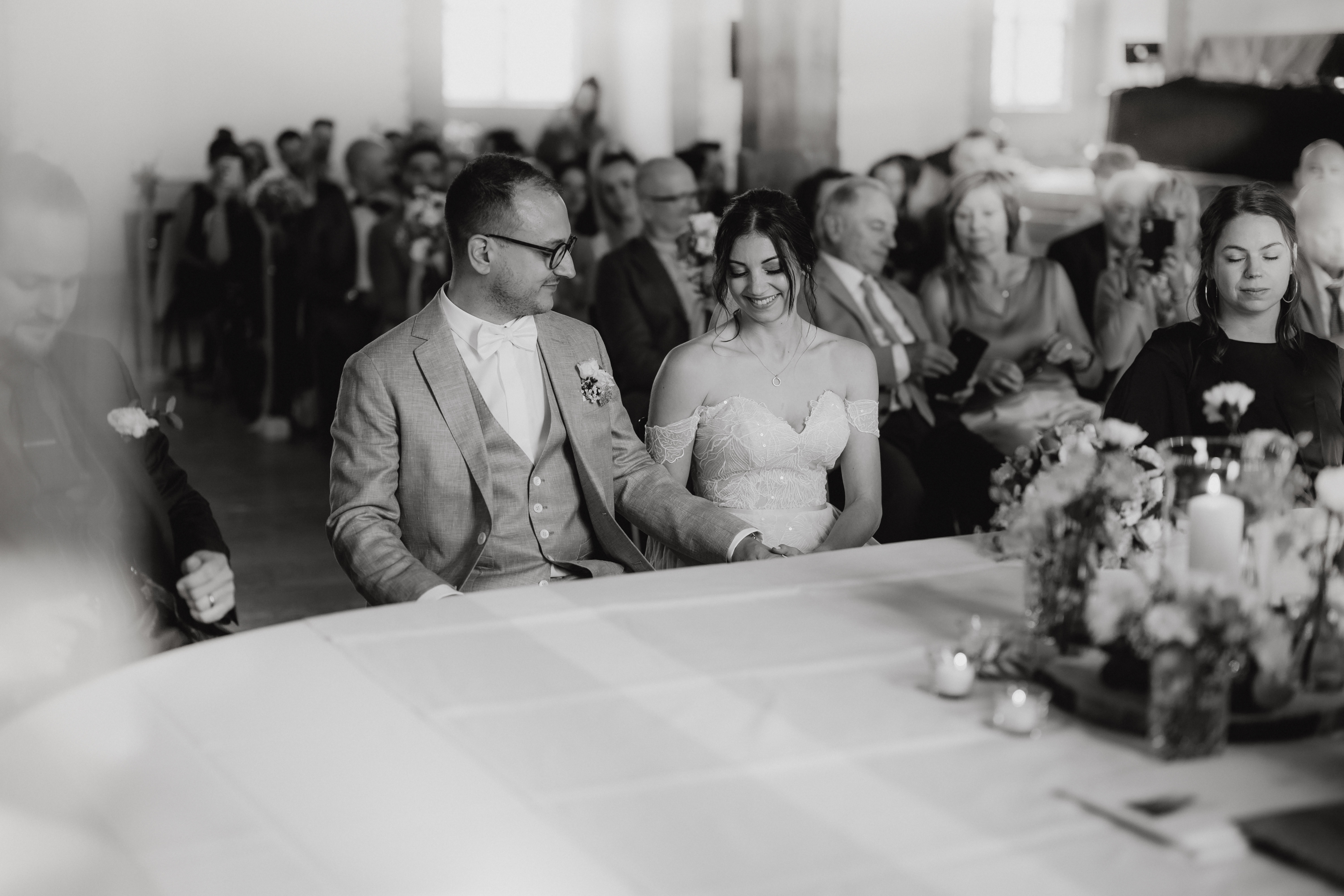 Schwarz-Weiß-Foto, das Braut und Bräutigam im Schloss Lenzburg zeigt, wie sie Händchen halten und lächeln. Der Bräutigam trägt einen hellen Anzug und eine Fliege, während die Braut in einem schulterfreien Brautkleid glänzt. Tasha Enders Hochzeitsfotografie fängt die Gäste ein, die im Hintergrund sitzen und zuschauen.
