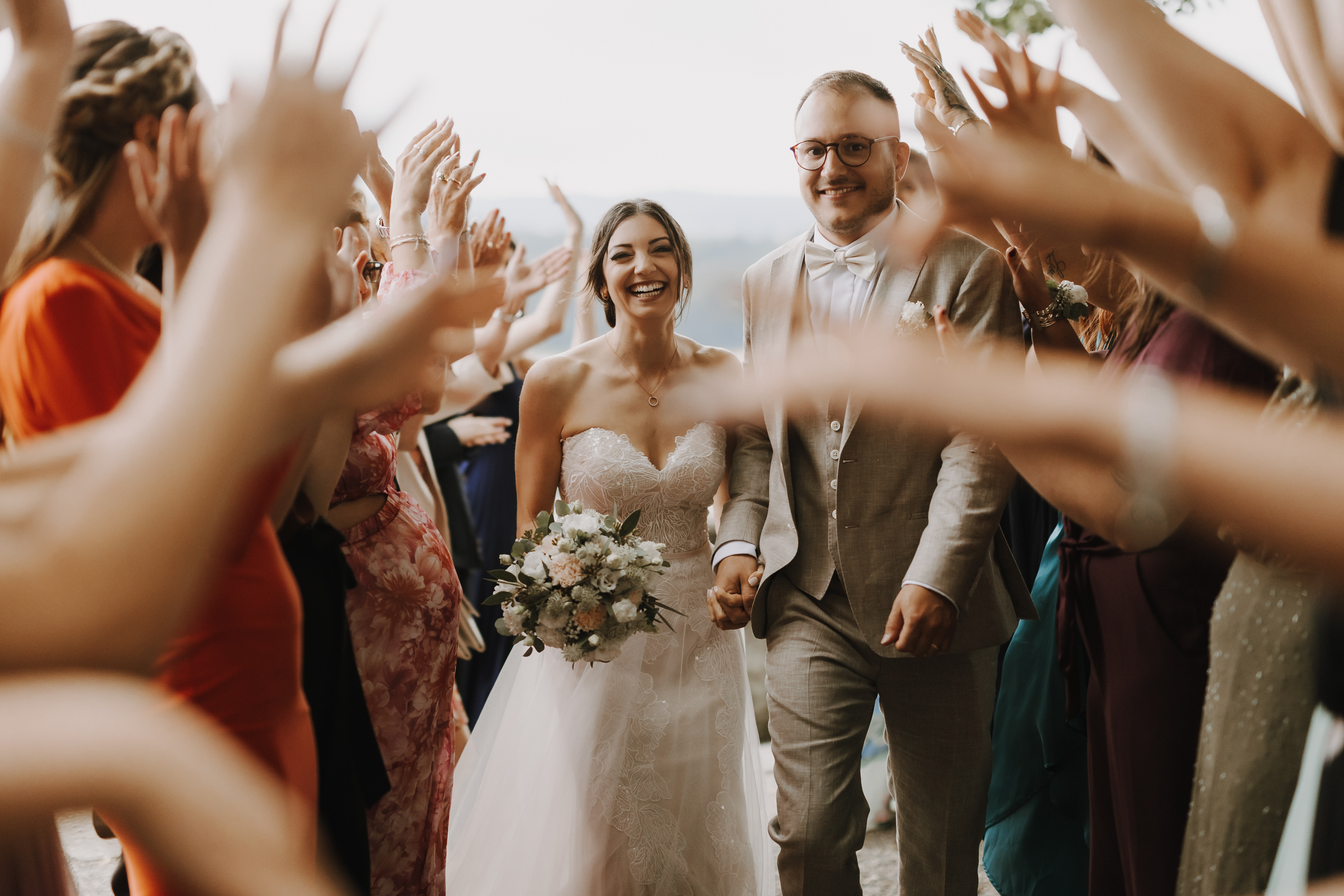 Eine Braut in einem trägerlosen weißen Kleid und ein Bräutigam in einem hellgrauen Anzug gehen lächelnd Hand in Hand, wunderschön eingefangen von *Hochzeitsfotografie* von Tasha Ender Photography. Sie sind von jubelnden Gästen umgeben, die ihre Hände zu einem Bogen erheben. Die Braut hält einen Blumenstrauß, mit einer verschwommenen Außenkulisse im Aargau als Hintergrund.