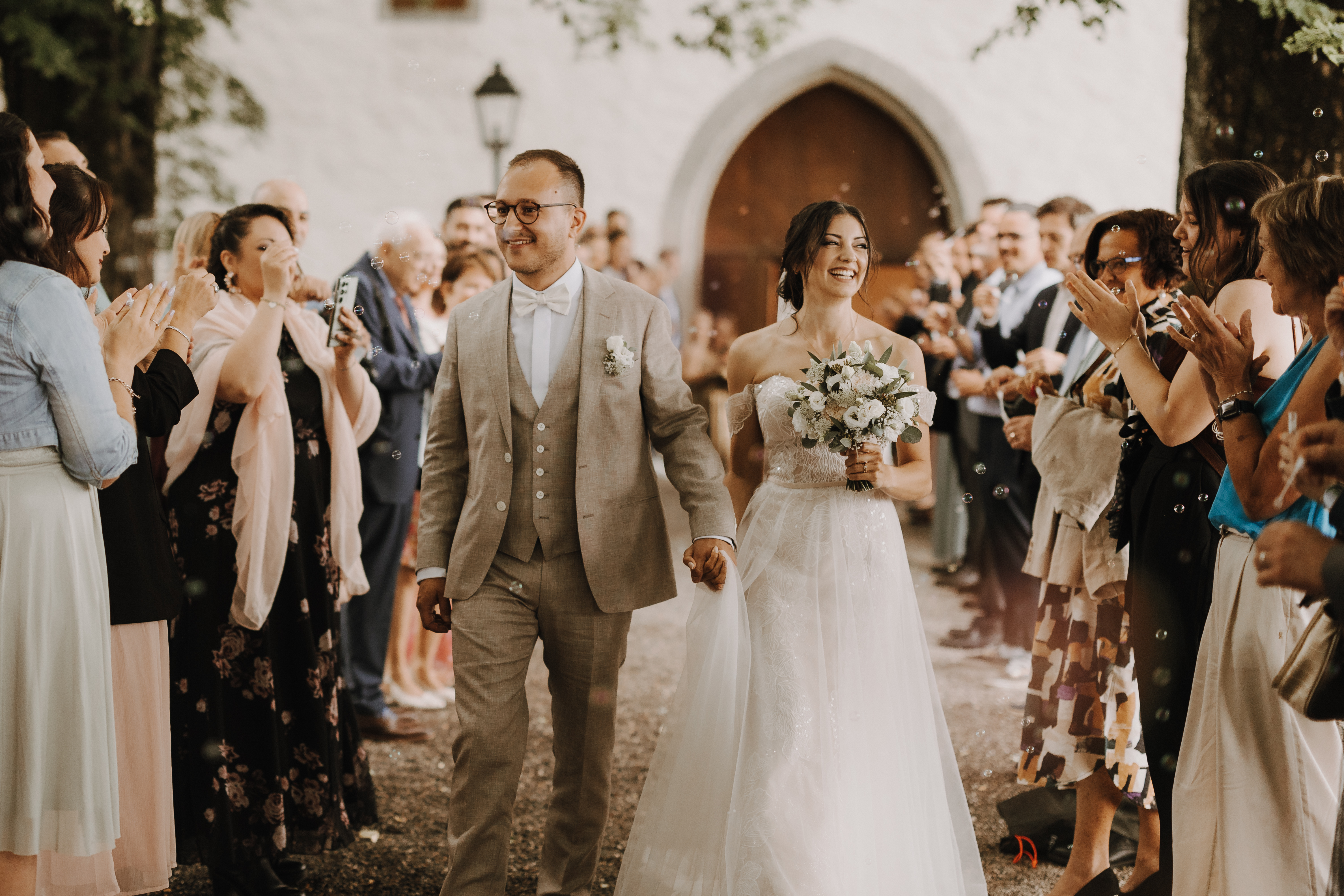 Braut und Bräutigam gehen Hand in Hand einen Gang im Freien in Baden entlang, gesäumt von applaudierenden Gästen. Der Bräutigam trägt einen beigen Anzug und eine Brille, während die Braut in einem weißen Spitzenkleid umwerfend aussieht und einen wunderschönen Blumenstrauß hält. Beide lächeln freudig und feiern ihren Hochzeitstag. Dieser wundervolle Moment wurde von Tasha Ender Photographys Hochzeitsfotografie-Service festgehalten.