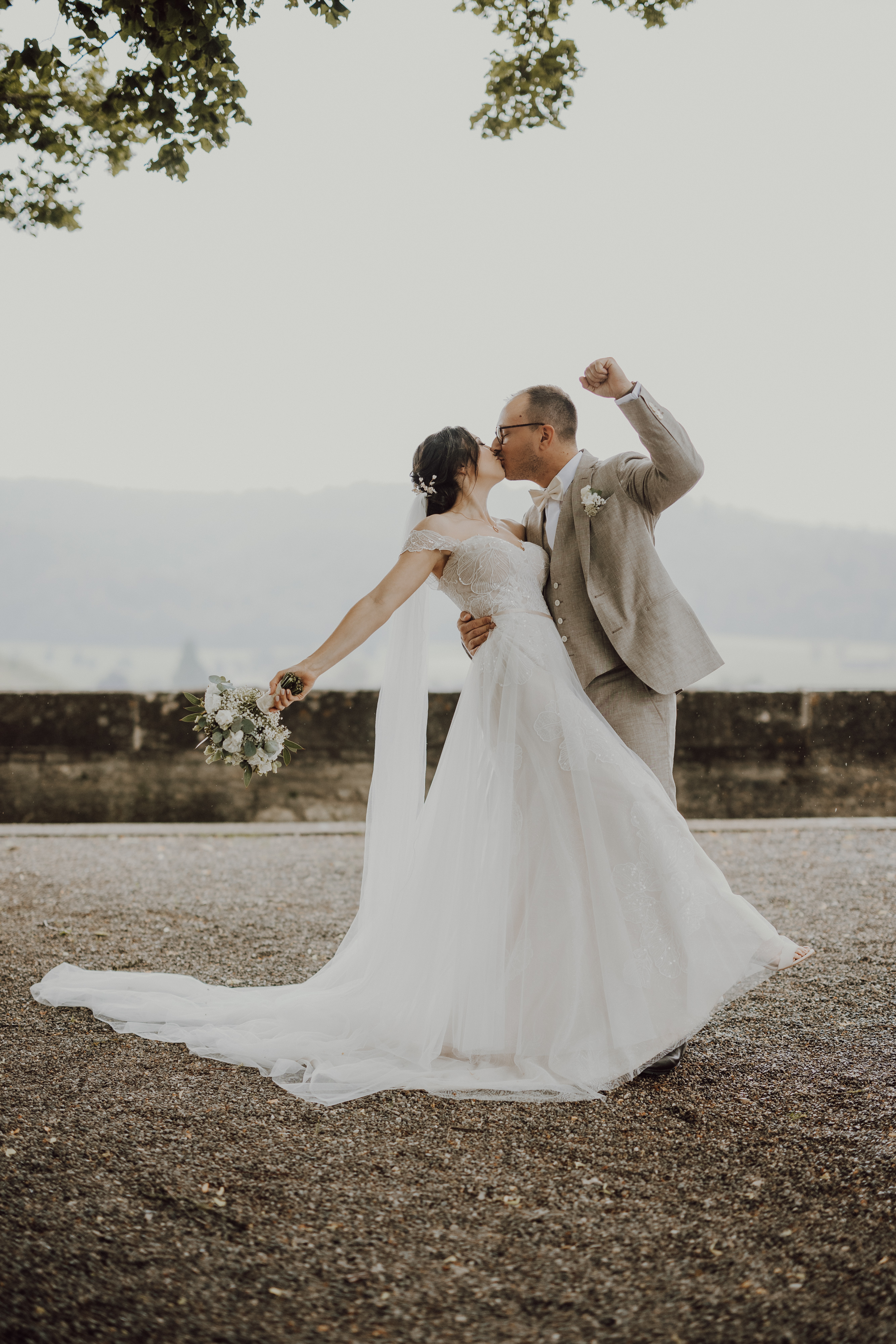 Ein frisch vermähltes Paar küsst sich leidenschaftlich im Freien in Baden. Die Braut, geschmückt mit einem fließenden weißen Kleid und einem Blumenstrauß in der Hand, lehnt sich in den Kuss. Der Bräutigam, gekleidet in einen hellgrauen Anzug, hebt triumphierend seinen Arm. Sie stehen auf einem Kiesweg mit Bäumen und dunstigen Hügeln im Hintergrund – eine exquisite Aufnahme aus der Hochzeitsfotografie-Kollektion von Tasha Ender Photography.