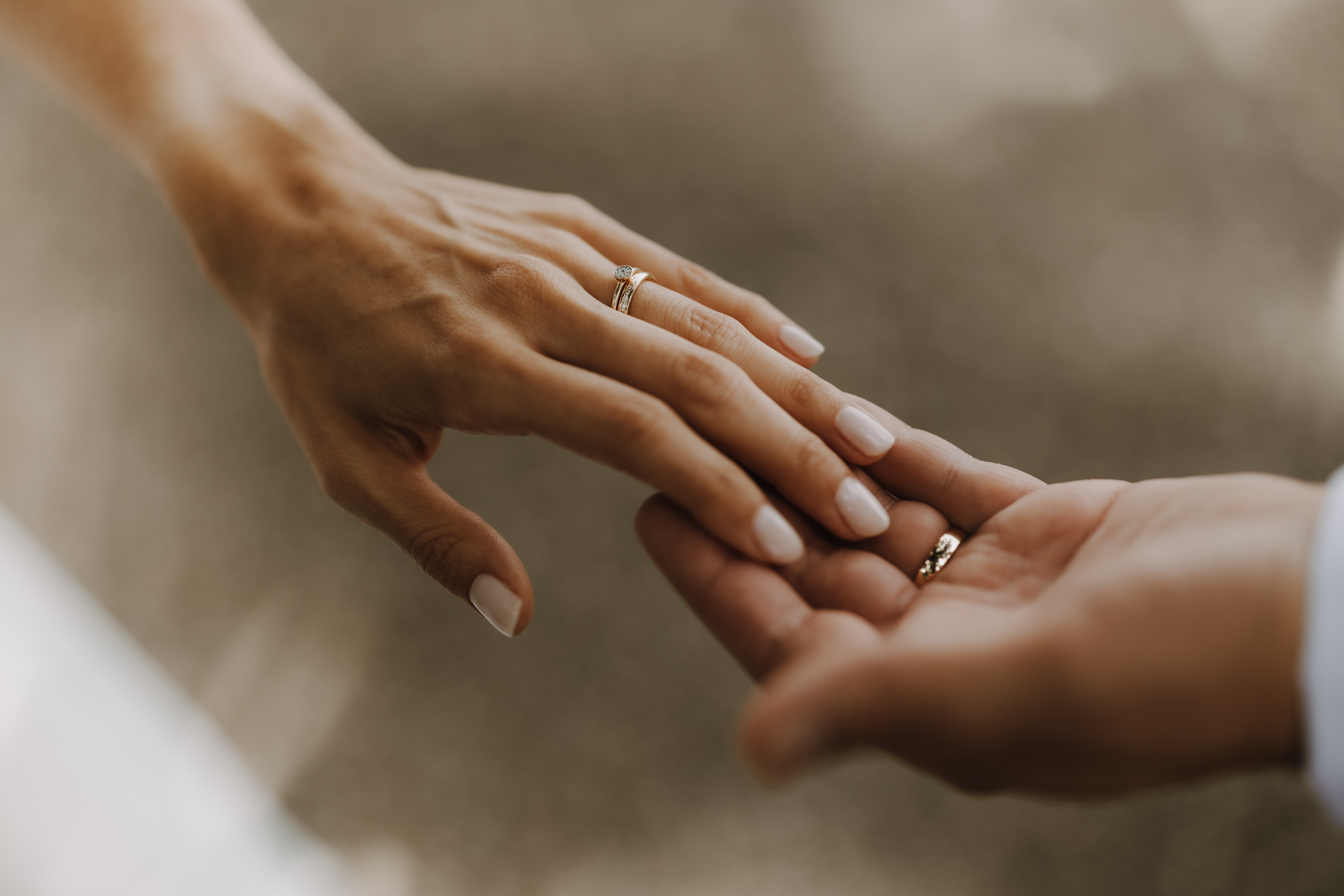Nahaufnahme zweier Hände, eine trägt einen Ehering, die andere bietet einen Ring an. Die Hand mit dem Ring hat helle Haut und manikürte Nägel, während die andere, leicht unscharf, einen Ring hält. Der Hintergrund ist leicht verschwommen – ein perfekter Moment von Tasha Ender, eingefangen während der Hochzeitsfotografie.