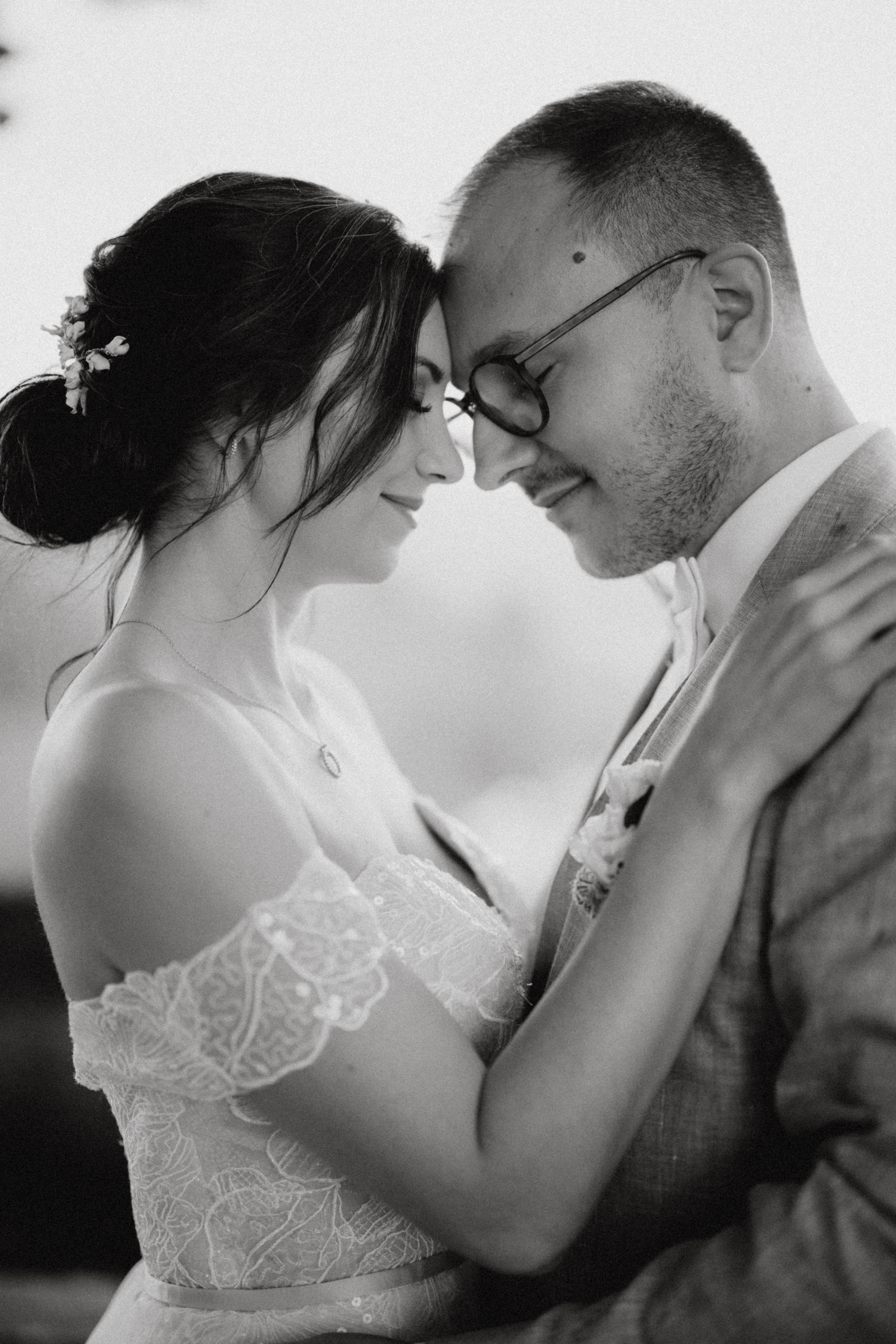 Mithilfe des Hochzeitsfotografie-Service von Tasha Ender Photography fängt ein Schwarzweißfoto, das von einem talentierten Fotografen in Zürich aufgenommen wurde, einen intimen Moment zwischen einer Braut in einem Spitzenkleid mit hochgestecktem Haar und einem Bräutigam mit Brille und Anzug ein. Sie stehen nah beieinander, ihre Stirnen berühren sich, die Arme umeinander geschlungen, die Augen geschlossen, und teilen diesen besonderen Moment.