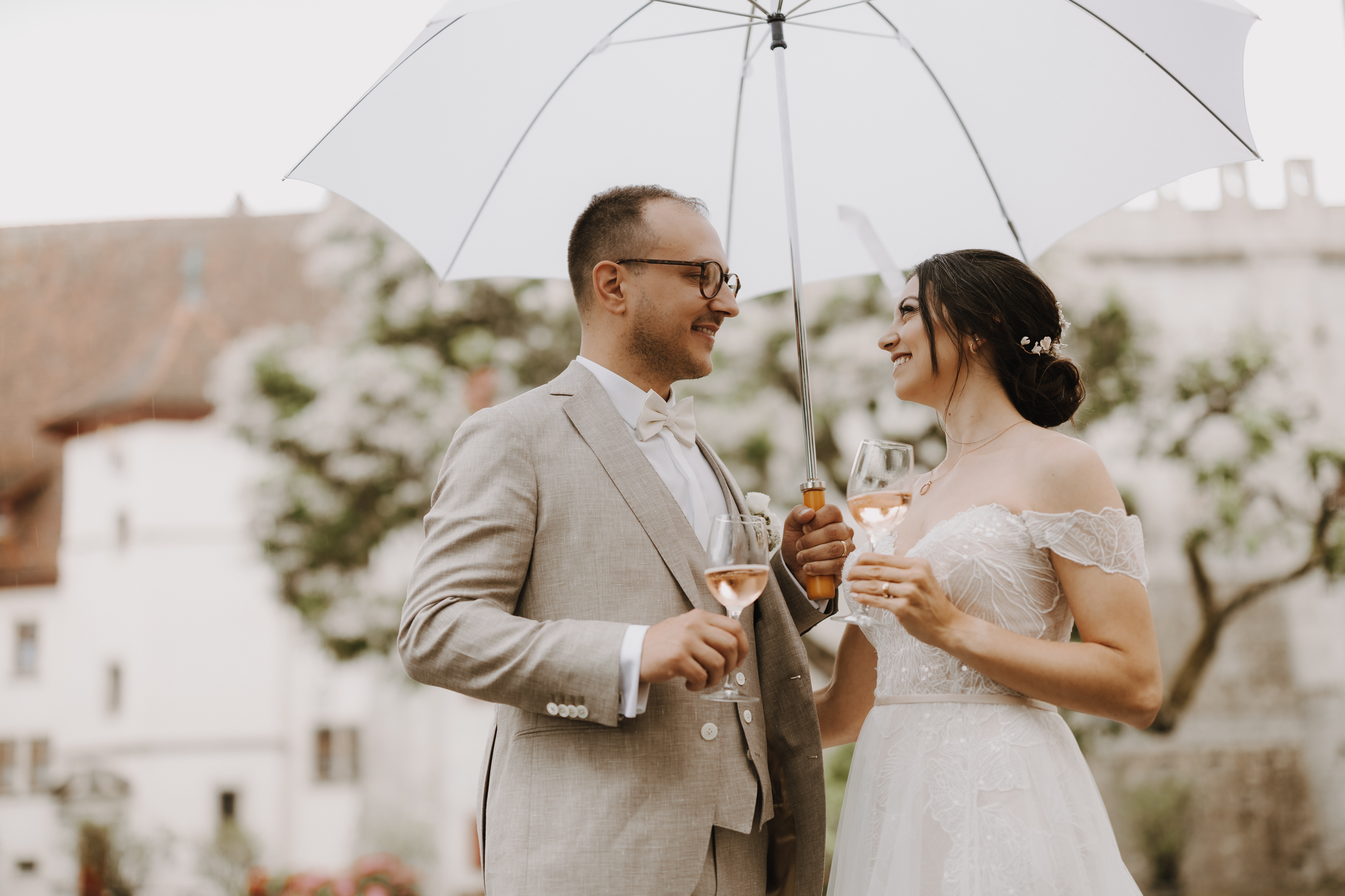Eine Braut in einem atemberaubenden weißen schulterfreien Kleid und ein Bräutigam in einem eleganten hellgrauen Anzug lachen fröhlich, während sie Weingläser halten und unter einem makellos weißen Sonnenschirm stehen. Die ruhige Kulisse fängt die malerische Schönheit von Baden ein, mit seinen üppigen Bäumen und der historischen Architektur, die den Charme des Augenblicks verstärken. Diese bezaubernde Szene wird von Tasha Ender Photography's Hochzeitsfotografie zum Leben erweckt.