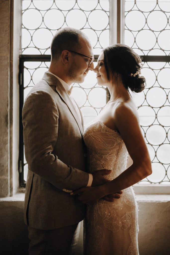 Ein Paar in Hochzeitskleidung steht eng beieinander vor einem rund gemusterten Fenster. Der Bräutigam trägt einen hellen Anzug und eine Brille, während die Braut ein trägerloses Spitzenkleid trägt und ihr Haar hochgesteckt und mit kleinen Blumen geschmückt ist. Sie blicken sich liebevoll an und fangen die Essenz der Hochzeitsfotografie ein.